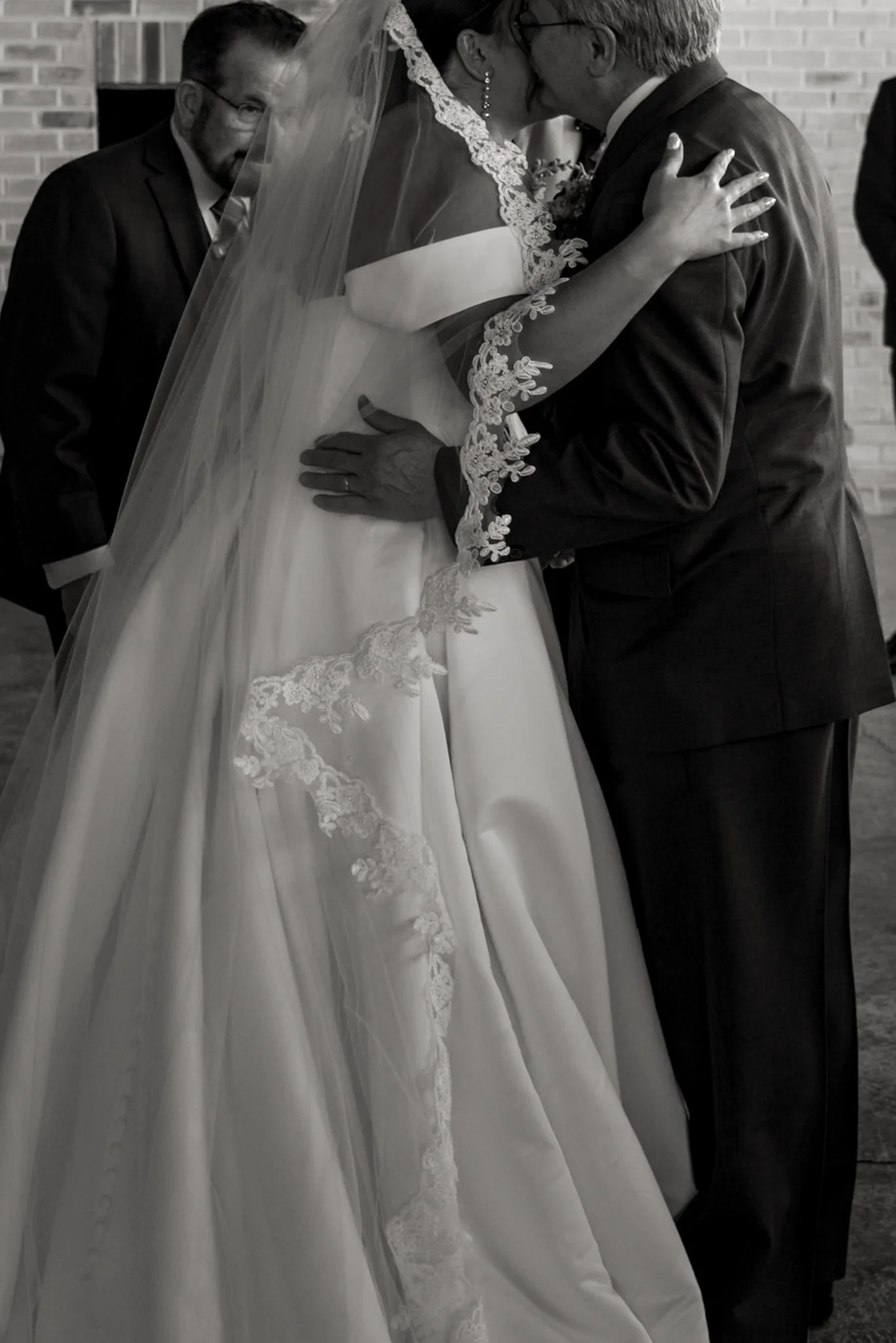 A bride and groom share a kiss at their wedding ceremony, with a man in a suit and glasses watching in the background. The bride is dressed in a wedding gown with lace details and a veil, and the groom is in a dark suit.