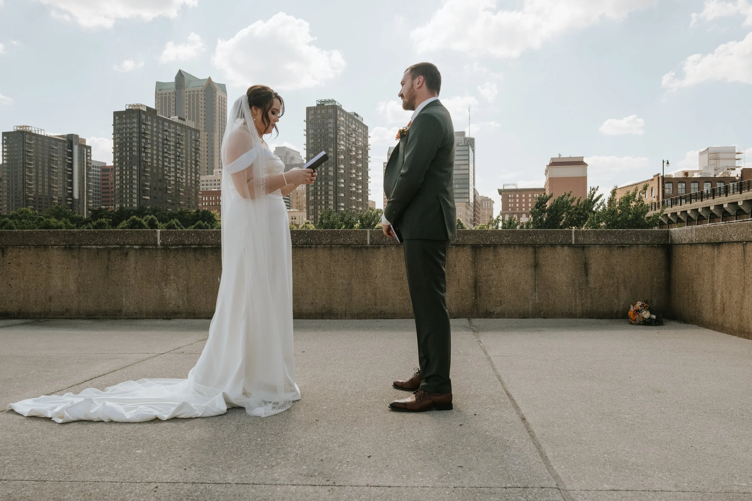 A bride and groom standing face to face outdoors in front of a city skyline, during a wedding ceremony. The bride is holding a small book or card and is dressed in a white wedding gown with an off-the-shoulder design and a veil. The groom is dressed 
