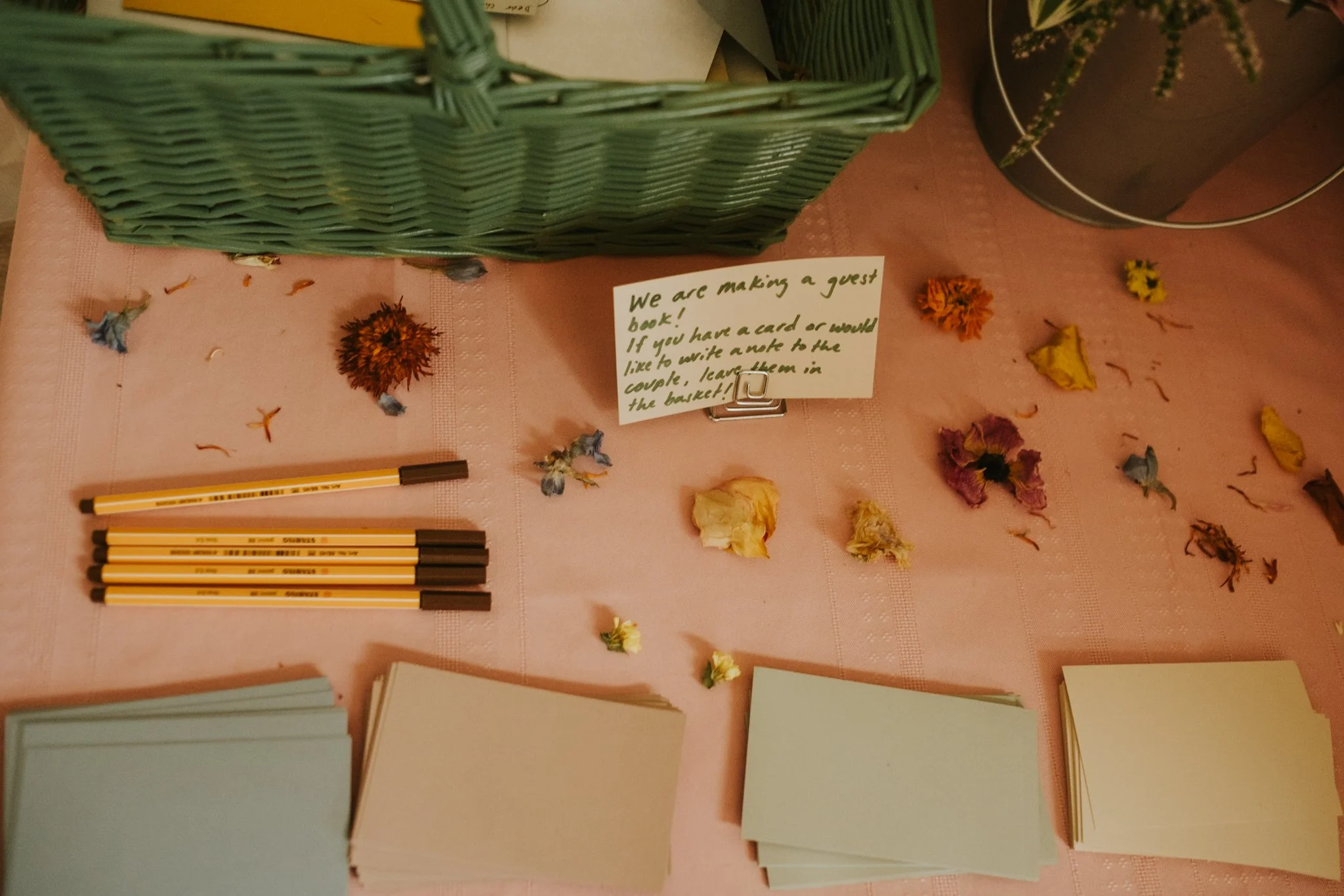 A pink tablecloth with scattered dried flowers, black and yellow markers at the bottom, a green basket, and a handwritten note on a clip suggesting making books and writing notes.