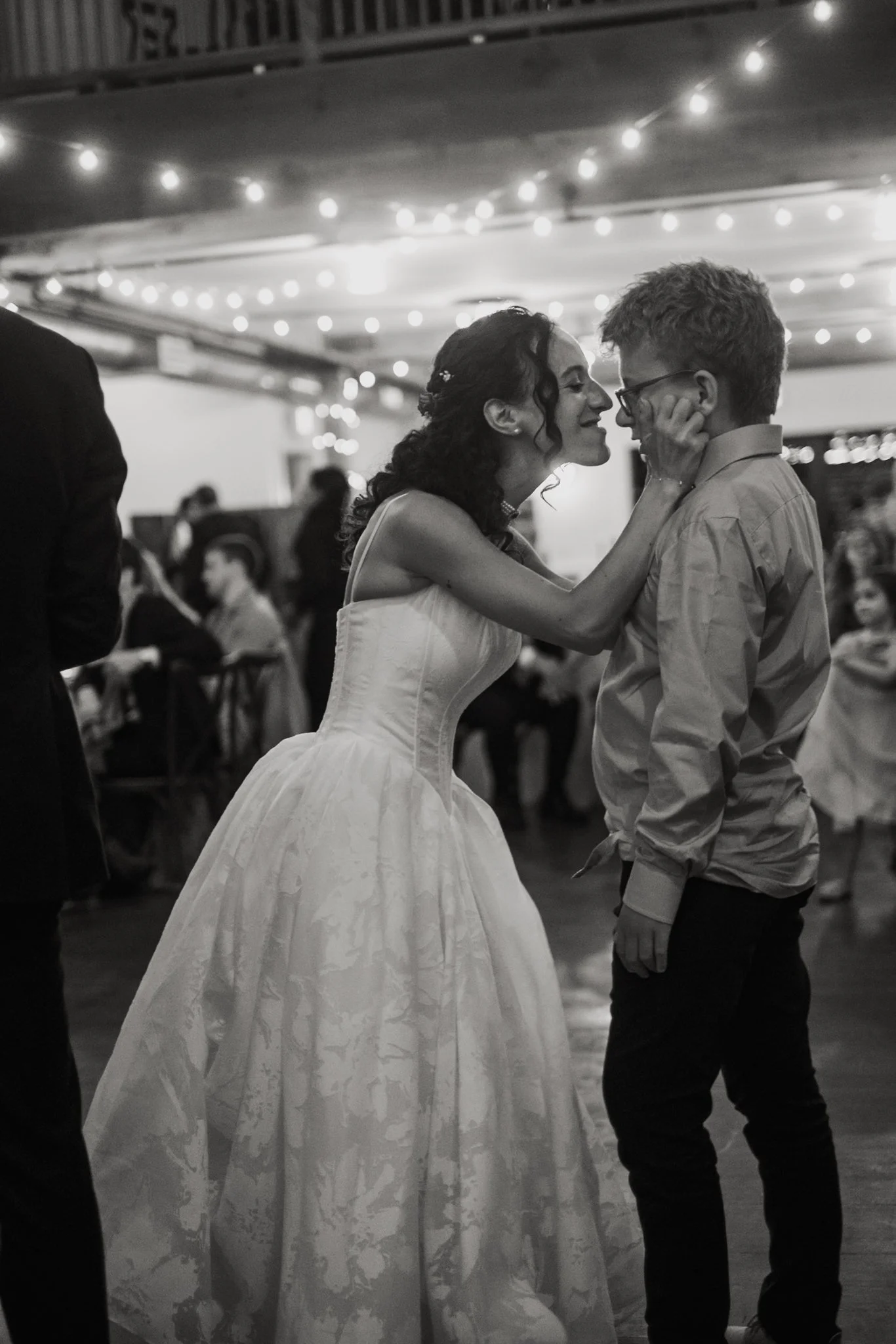 Black and white photo of a woman and man dancing closely at an indoor event, with festive string lights overhead and people in the background.