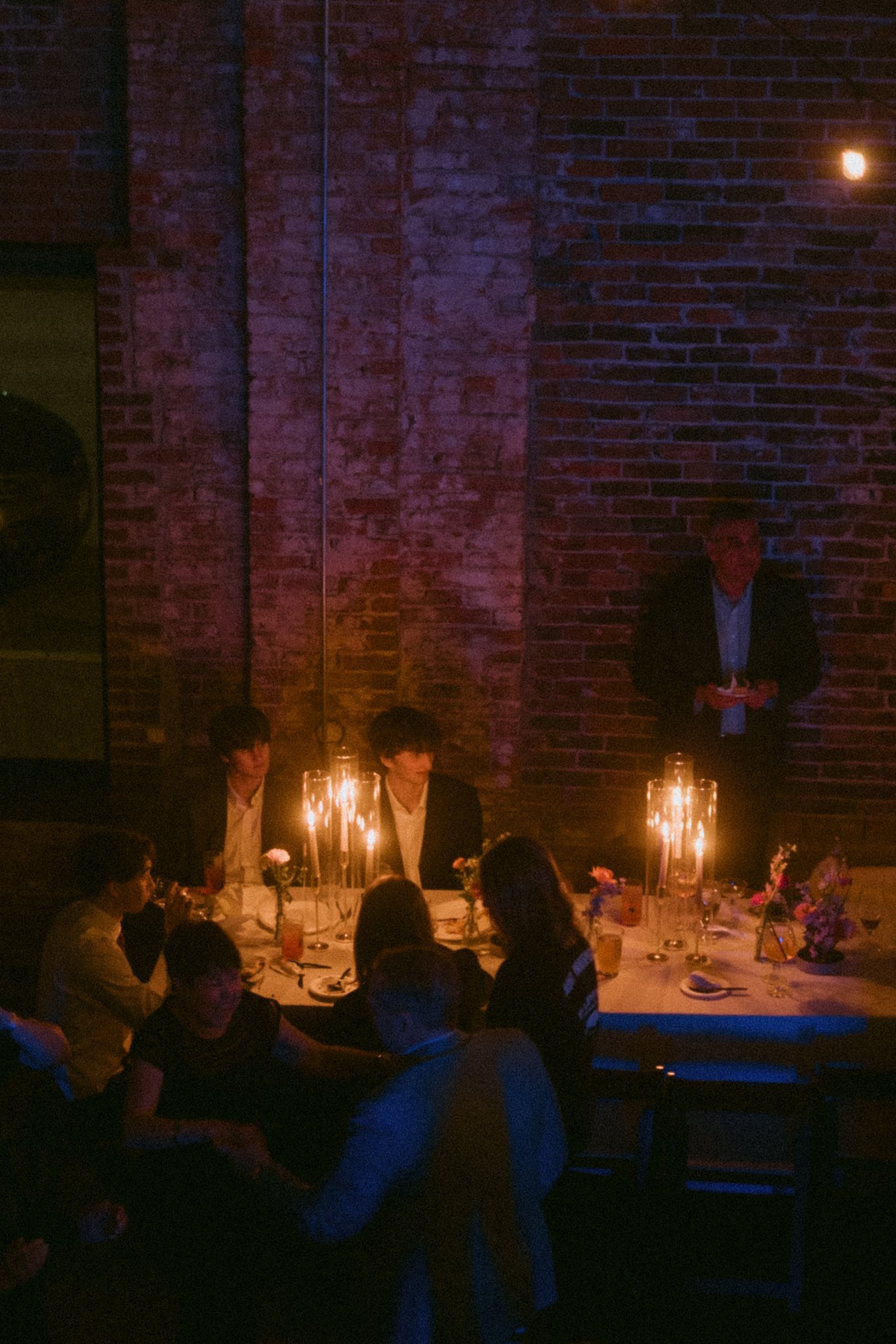 People sitting around a round table in a dimly lit restaurant, with at least one man standing in front of a brick wall, possibly giving a speech or toast. The table is decorated with candles and flowers.