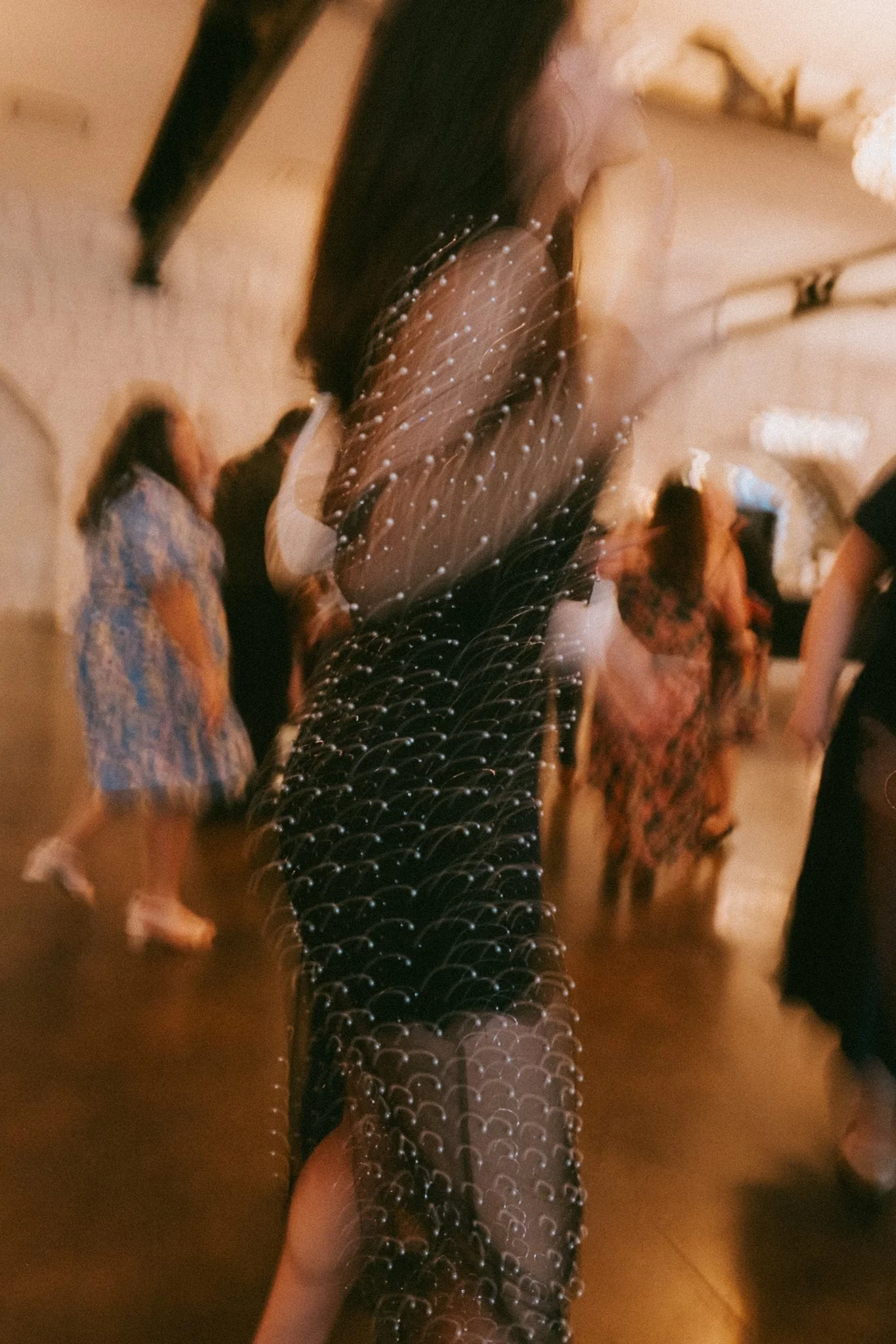 A woman in a sheer black dress with shiny embellishments, standing in a room with people. The photo is taken in a blurry, warm-toned setting.