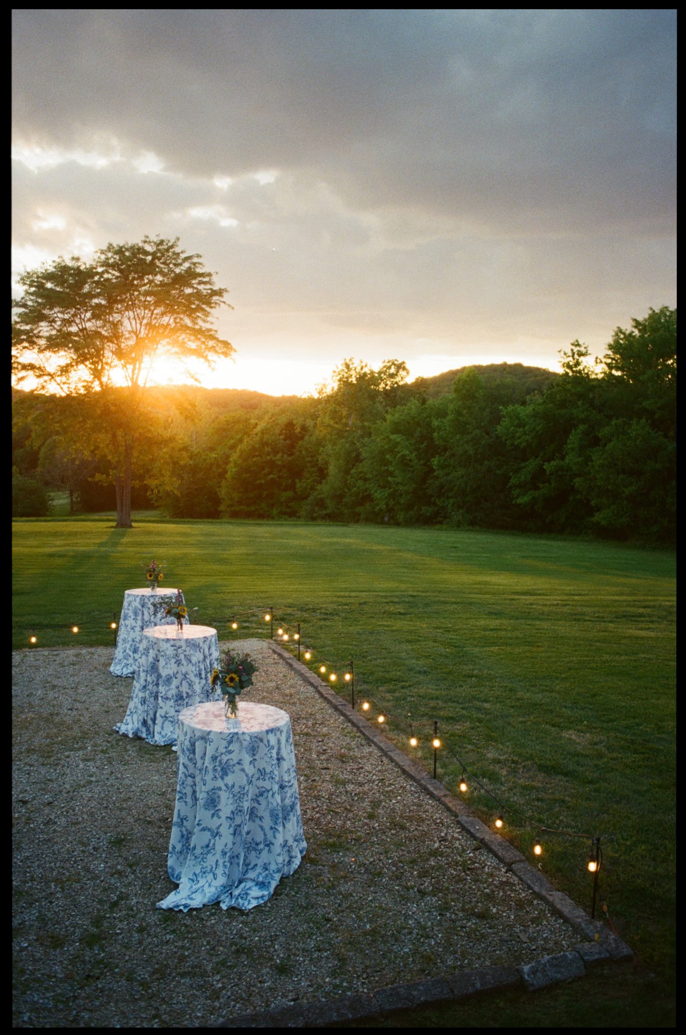 Sunset over a green lawn with trees in the background, decorated tables with blue floral tablecloths and small flower arrangements, string lights along the edge of the gravel area.