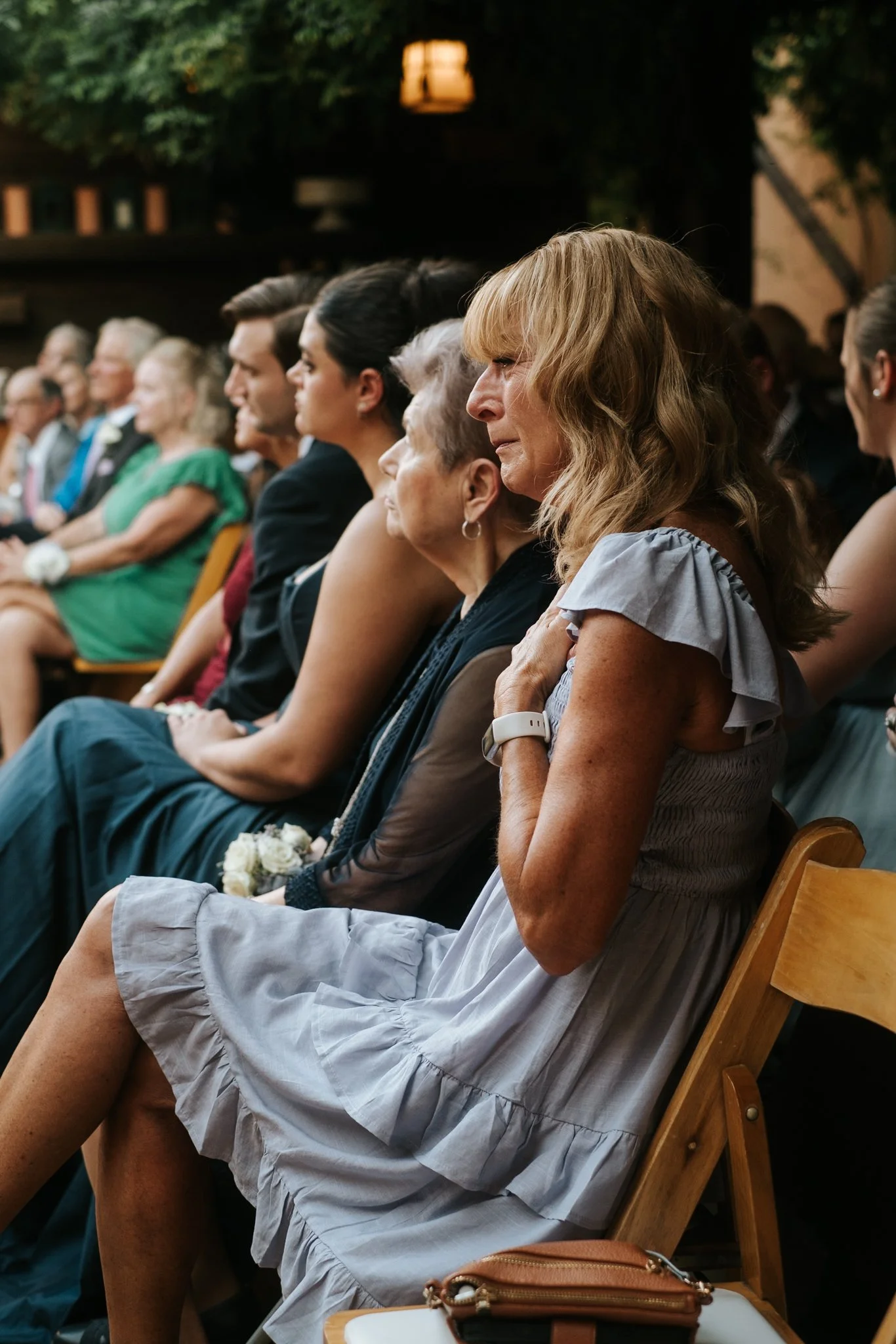 A line of people sitting on wooden chairs at an outdoor event, some with flowers, and a woman in a light gray dress, with blonde hair, sitting with her hands over her chest, looking serious or emotional.