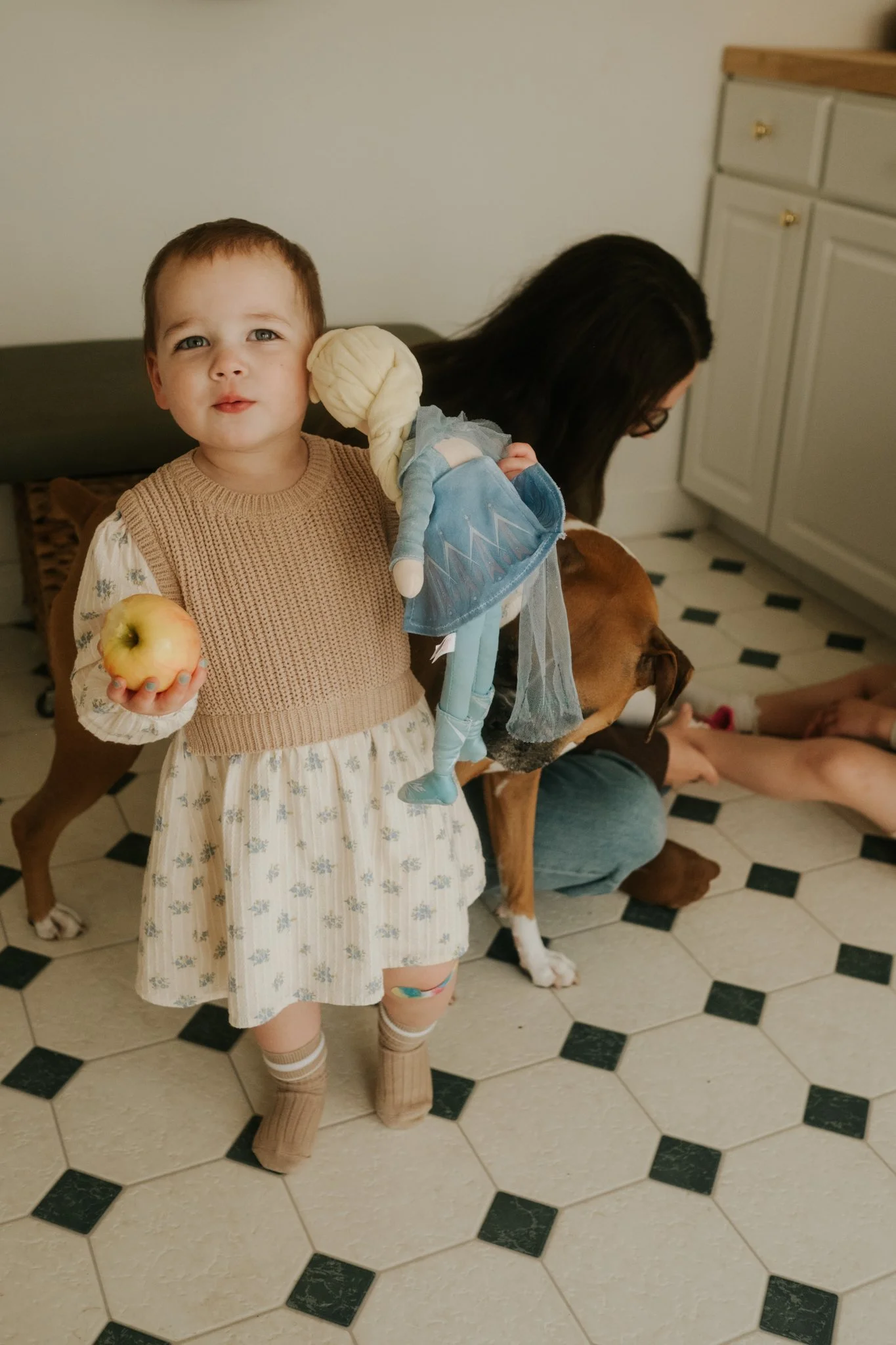 A young girl with short brown hair and light skin, wearing a beige knitted vest and a floral dress, standing on a black and white tiled floor. She holds an apple in her left hand and a doll over her shoulder. Behind her, a person with long dark hair,