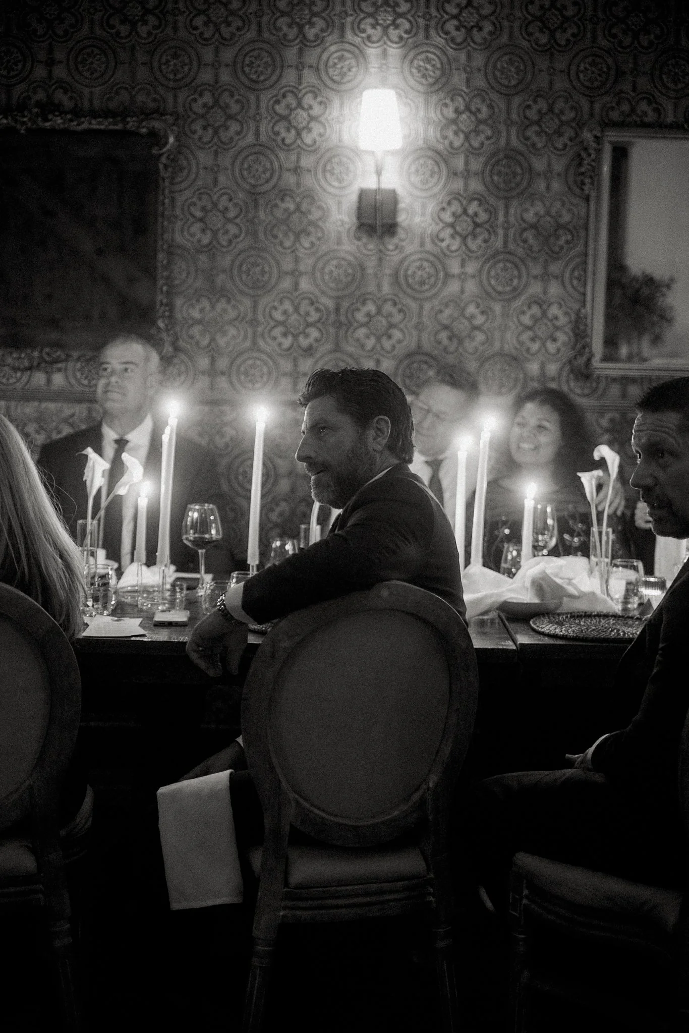 People sitting around a dining table in a fancy restaurant, with lit candles and wine glasses, against a patterned wall with a wall lamp.