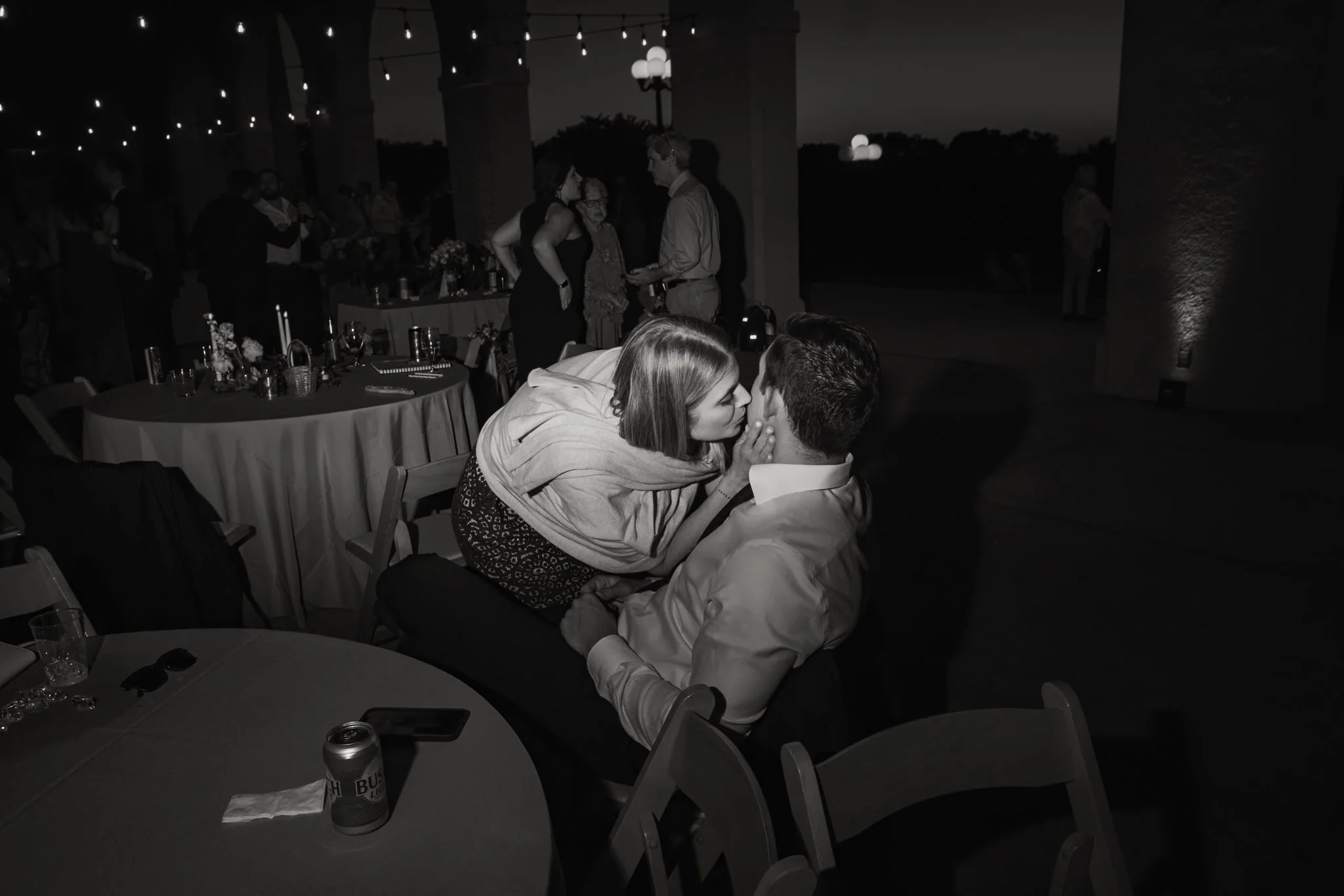 A night-time gathering of people at a celebratory event, with string lights overhead, people dancing and mingling, and a couple sharing a close moment at a table in the foreground.
