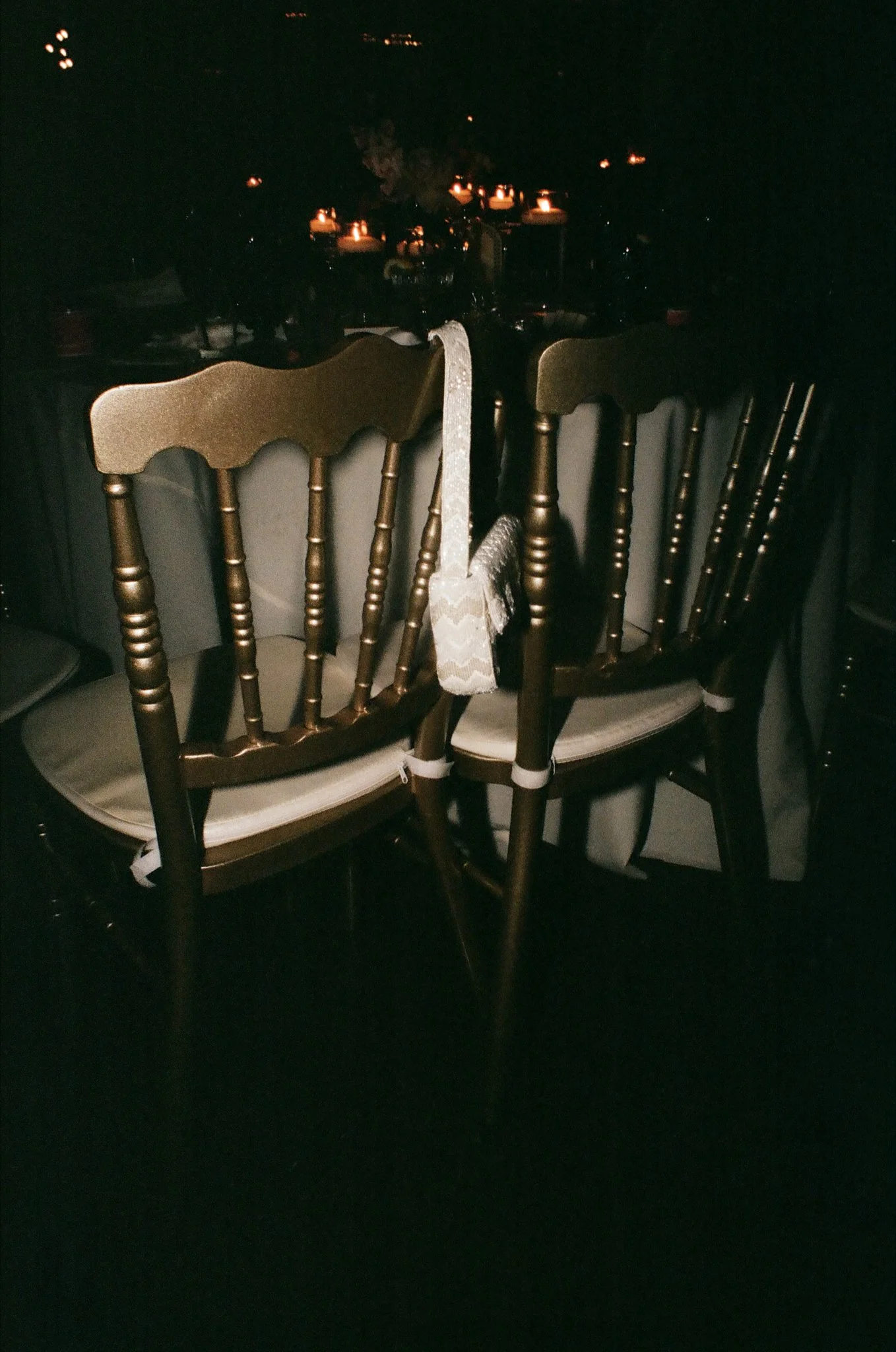 Dark dining room with a table set for a meal, chairs around the table, candles providing ambient lighting, and a lace cloth hanging from one chair.