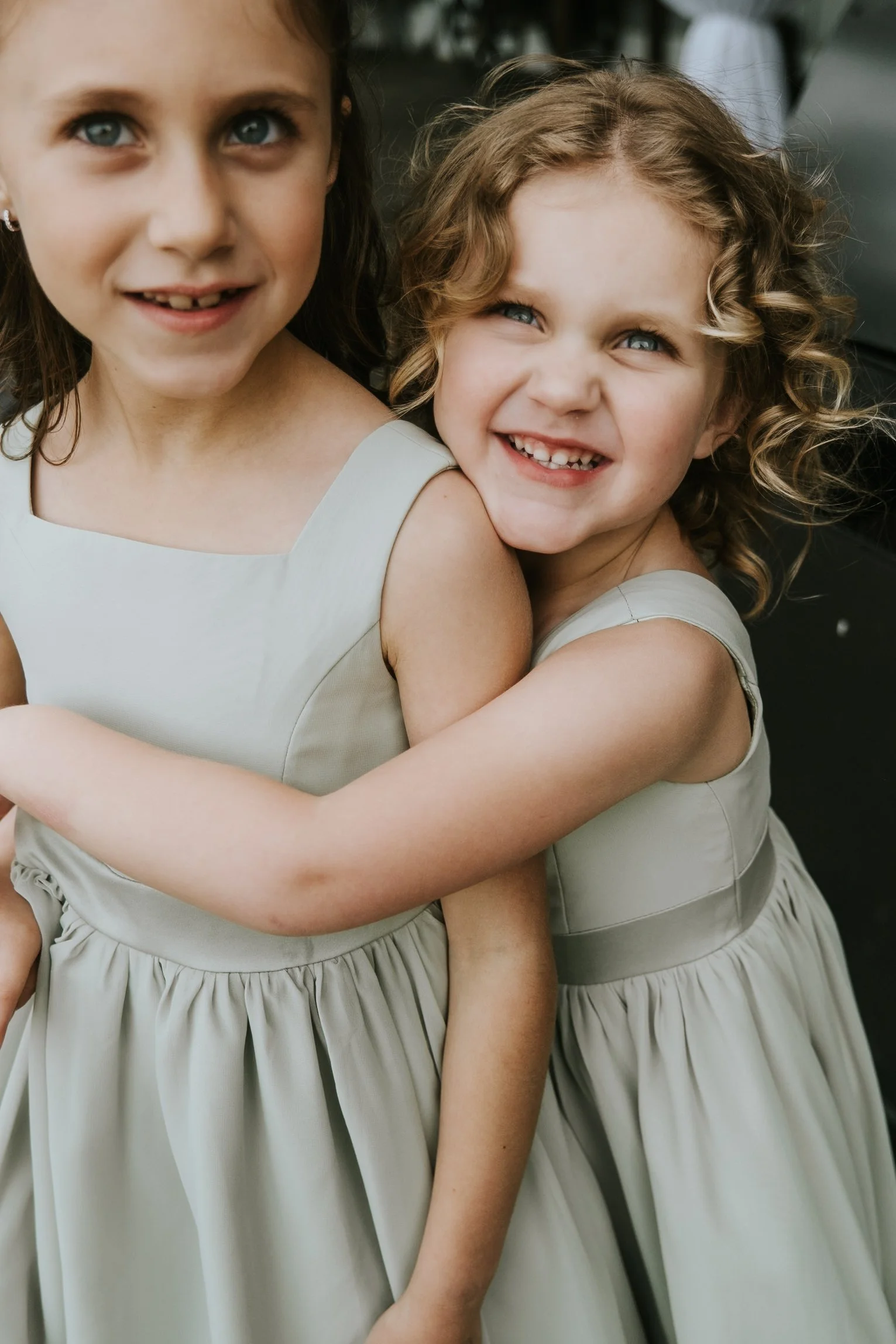 Two young girls wearing matching light-colored dresses hugging each other and smiling.