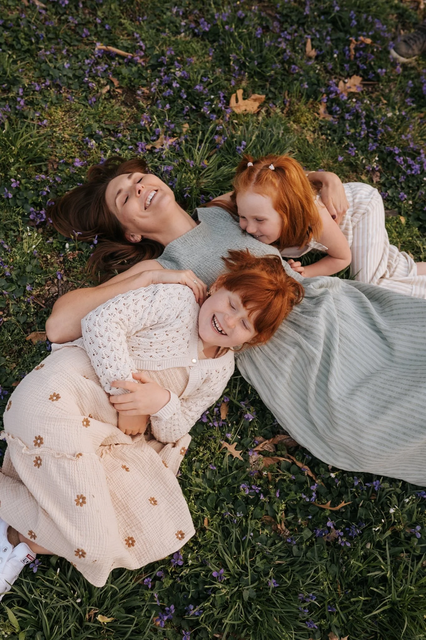 A woman with brown hair lies on her back in a field of purple flowers, with two young red-haired girls laying on her, all smiling and laughing.