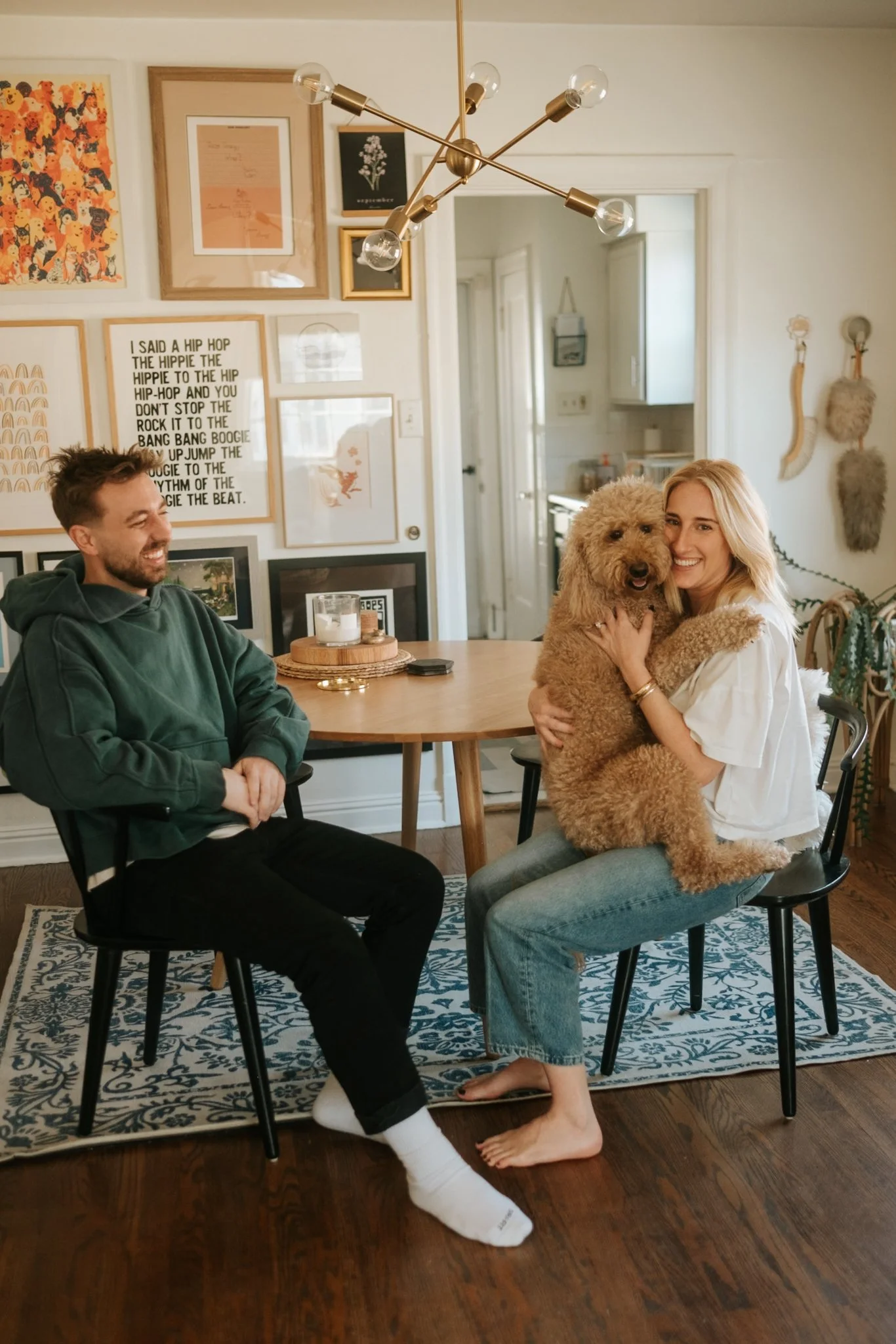 A woman with blonde hair holding a fluffy brown dog sitting on her lap, smiling. A man with brown hair wearing a green hoodie sitting nearby in a living room decorated with framed artwork, wall hooks, and a gold chandelier.