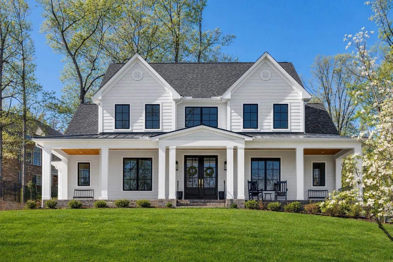White two-story house with black doors and windows, front porch with columns and benches, surrounded by green lawn and trees.