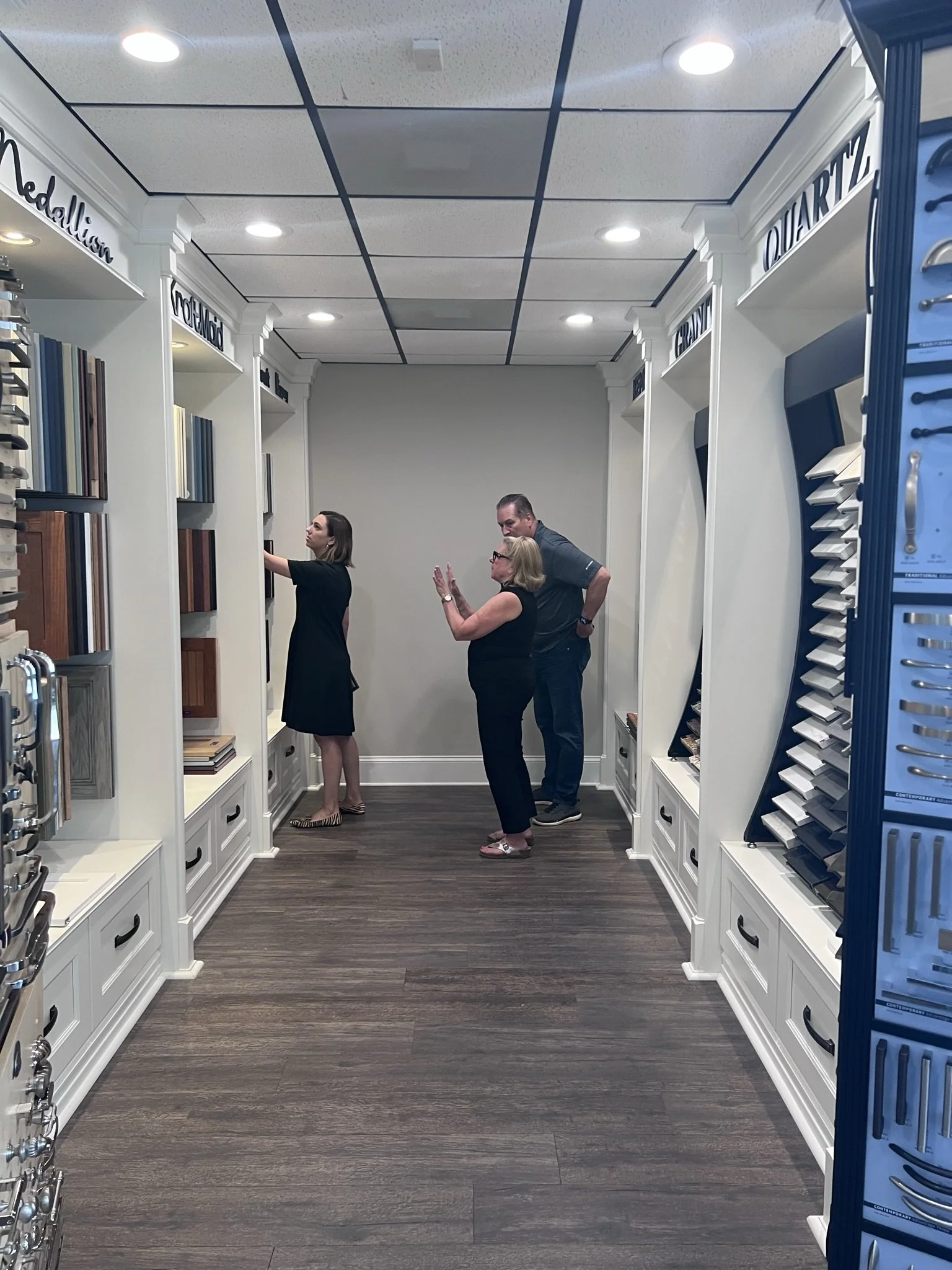 Three people inside a store, looking at display samples of wood and laminate flooring along white shelves.