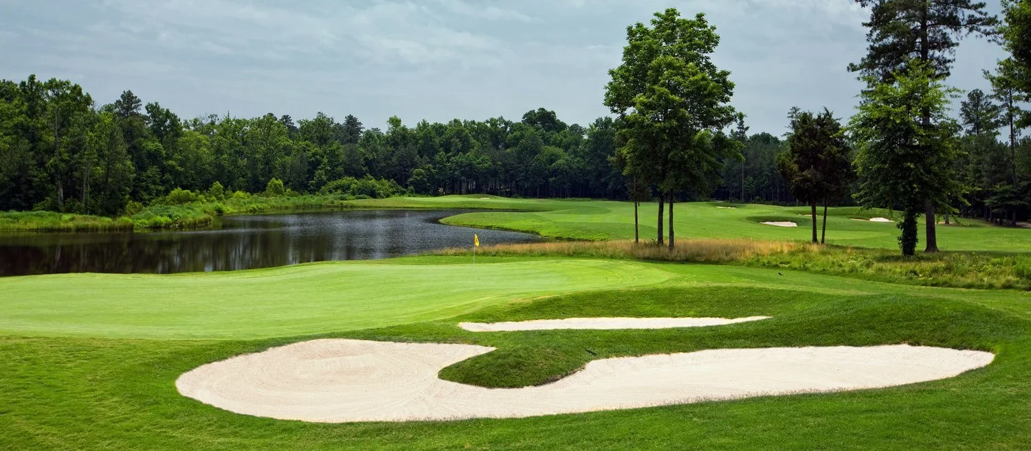 Golf course with green fairways, sand traps, and a water hazard surrounded by trees.