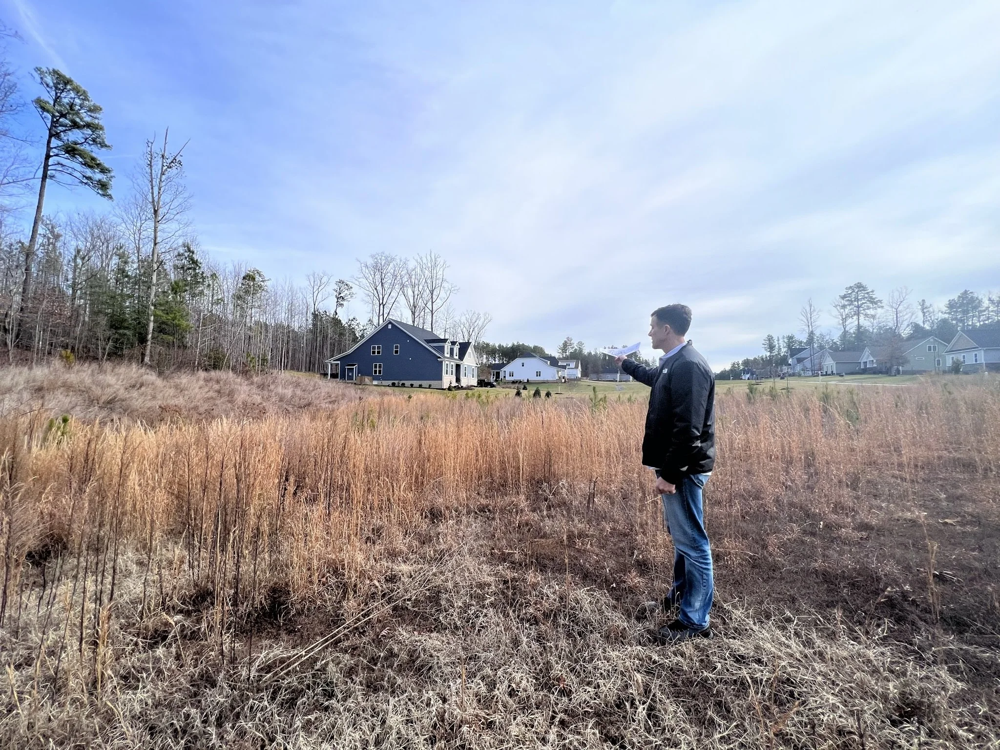 A man in a black jacket and jeans standing in a field of dry grass, pointing towards houses in the distance under a clear blue sky.
