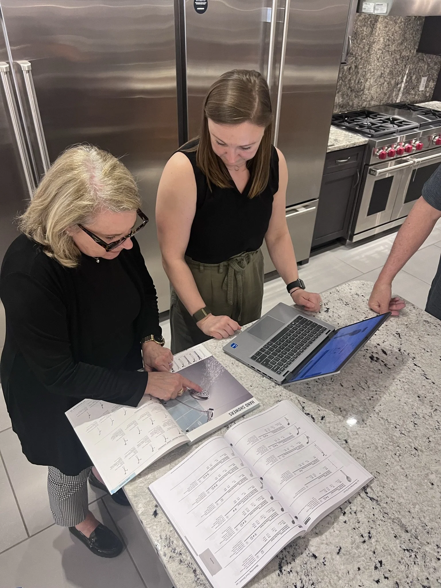 Two women are reviewing and discussing a cookbook and a laptop on a kitchen countertop, with a stainless steel refrigerator and stove in the background.