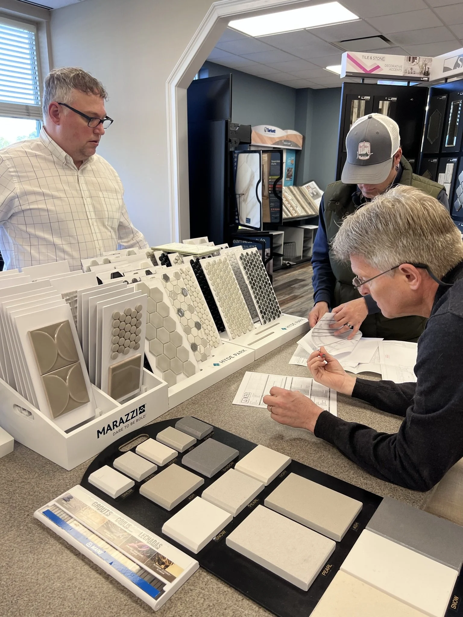 Three men examining tile samples in a showroom, with various tile samples displayed on a counter.