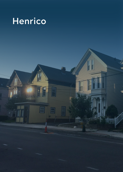 A row of houses on a street at dusk, with one house illuminated by a streetlight. The sky is clear and the word 'Henrico' is in the top left corner.