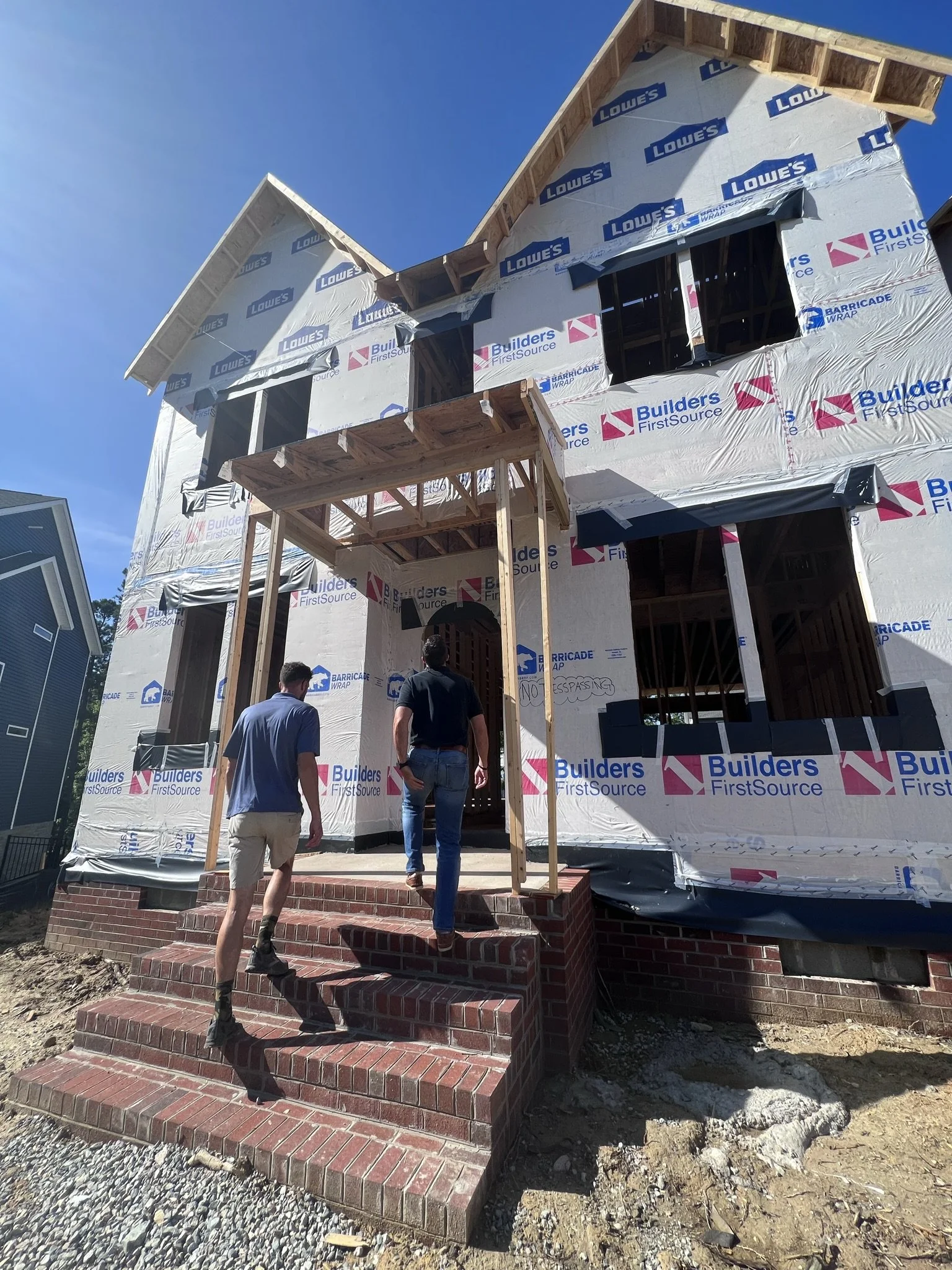 House under construction with two men walking up brick stairs towards the front door, wrapped in building materials, with a partly completed second story and a blue sky in the background.