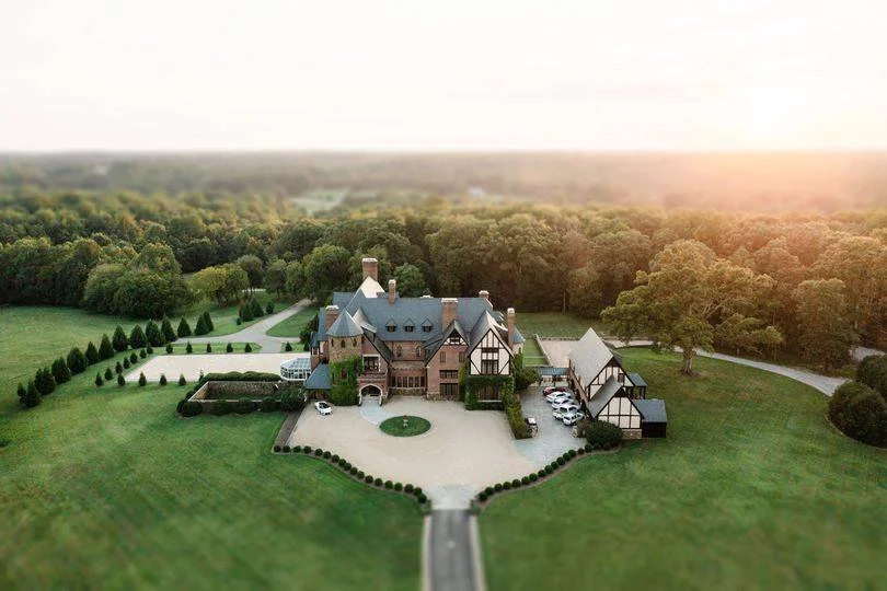 Large mansion with a driveway, surrounded by green lawns, trees, and landscaped gardens, set in a rural area.