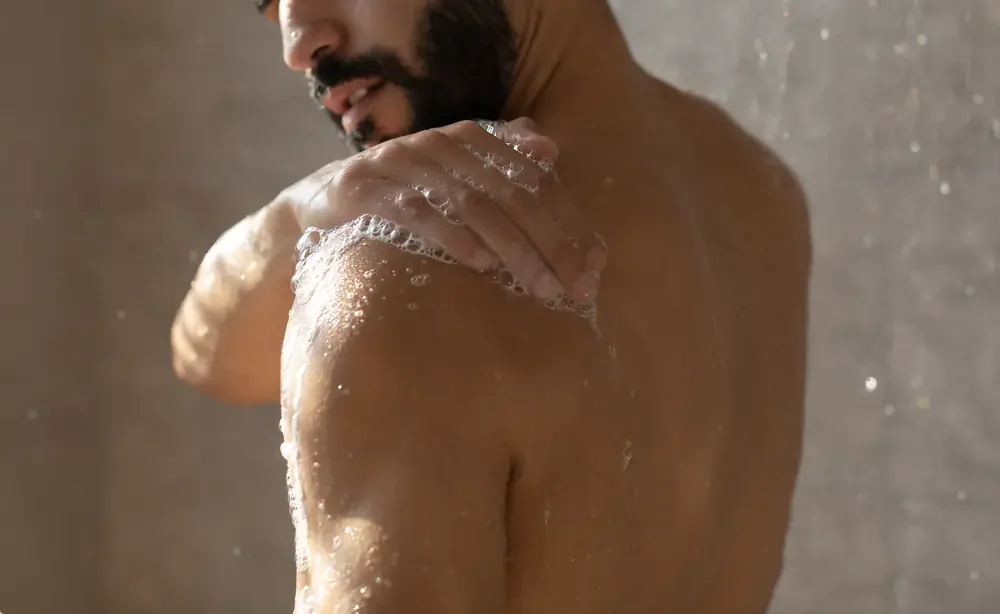 A man with a beard taking a shower, with water and soap suds on his shoulder and hand.