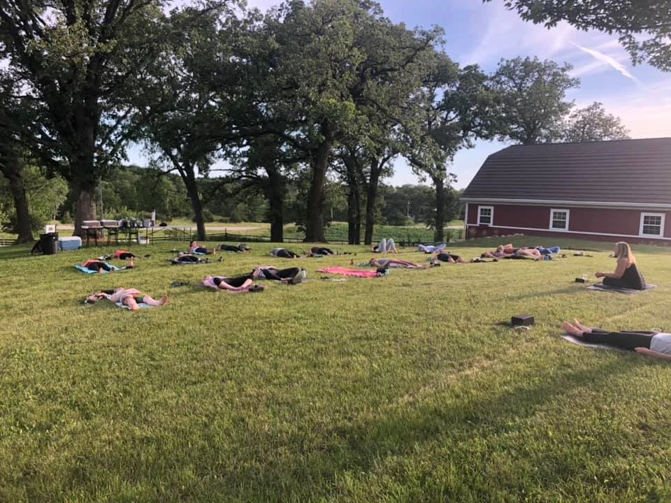 Yoga session at Twin Vines in Iowa. Shows a group over 15+ women laying down on yoga mats in the grass near trees with a red barn in background.