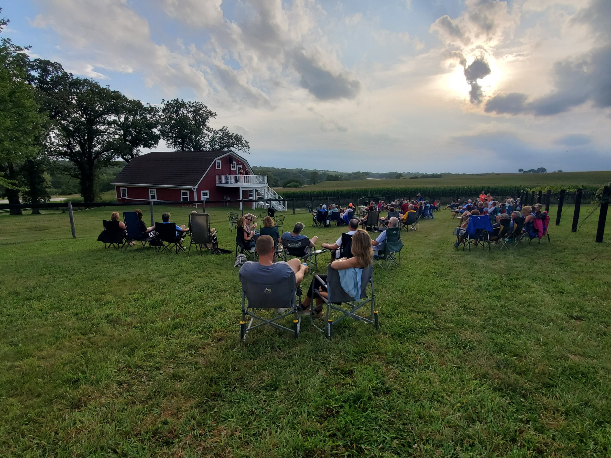 Live music event at Twin Vines. 25+ community members and families spread out on green lawn in foldable lawn chairs. Setting Sun and red barn venue in background.