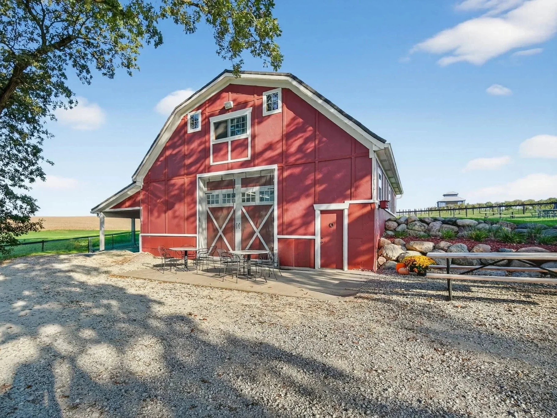 Red barn with white trim on a farm, with outdoor seating and a gravel area in front, surrounded by trees and fields, under a partly cloudy sky.