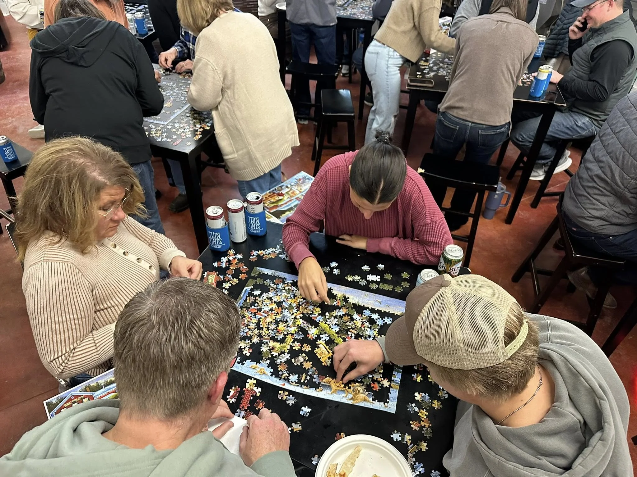 2025 holiday puzzle event at Twin Vines in Panora, IA. Image shows family of four working on a puzzle with four other families visible in the background at separate tables. Puzzle pieces, seltzers, beers, and pizza crust can all be seen on the table.