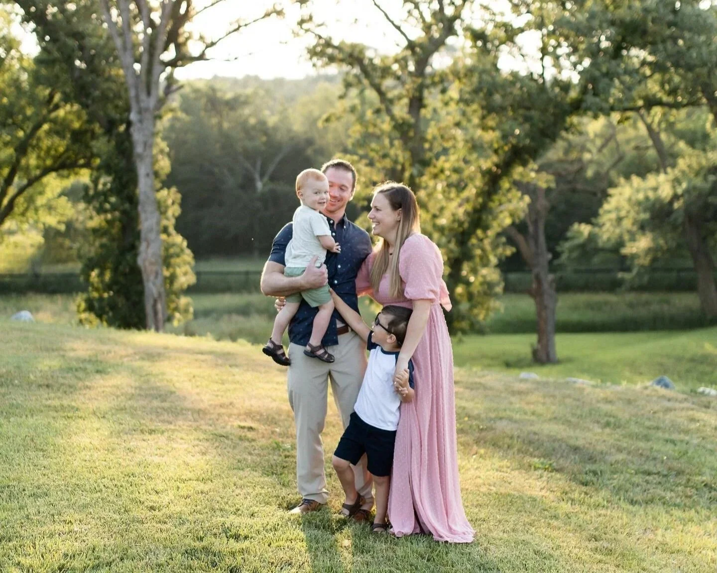 Family of four standing on grassy field with trees in the background, smiling and playing together during sunset.