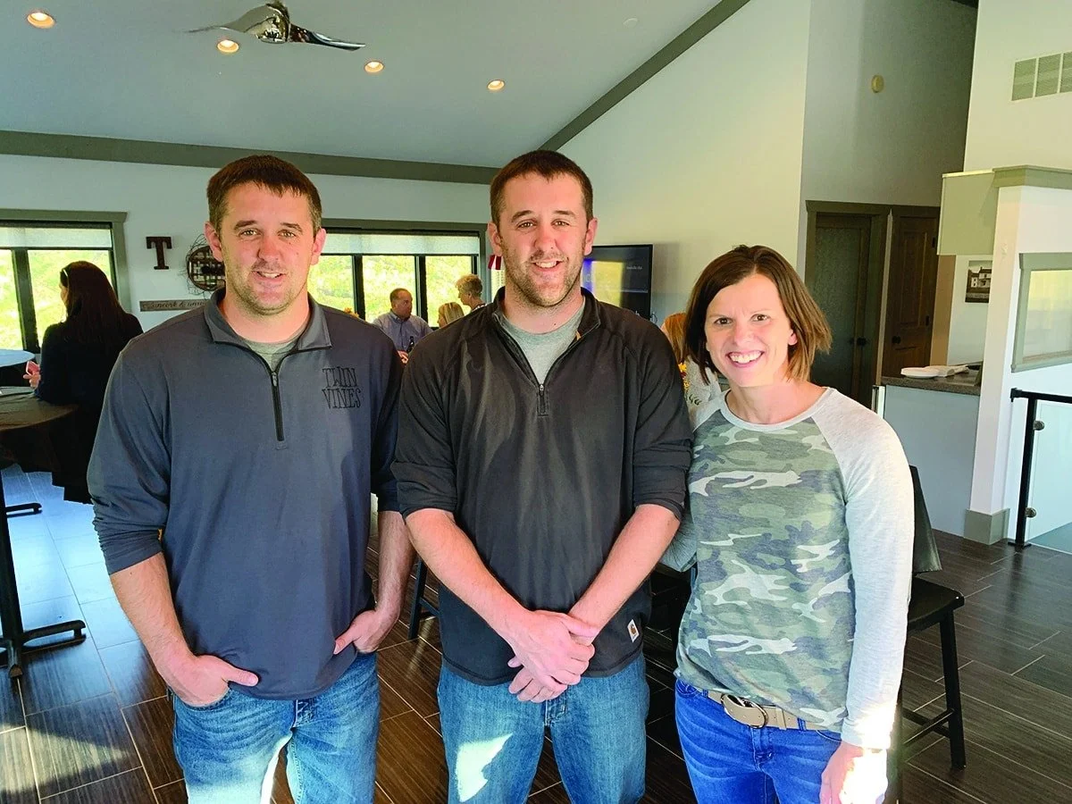 Twin Vines owners Ben and Brad Hayes smiling side by side inside the venue's main building. Wife beside brother on right, dark wood floors, bright walls with sage green trim, and group of customers behind the group of three.