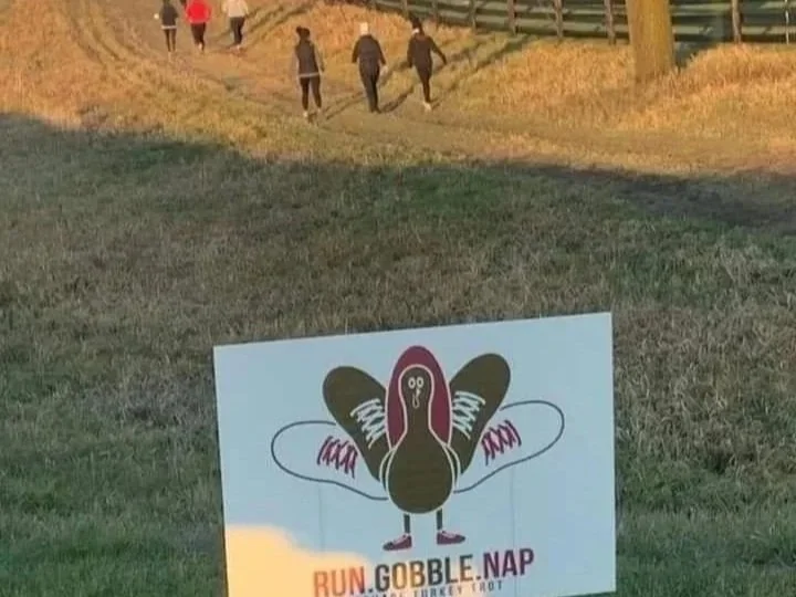 Participants running in the background past Turkey Trot signs at the annual Thanksgiving Turkey Trot hosted by Rephra E-Fitness at Twin Vines Vineyard in Panora, Iowa.
