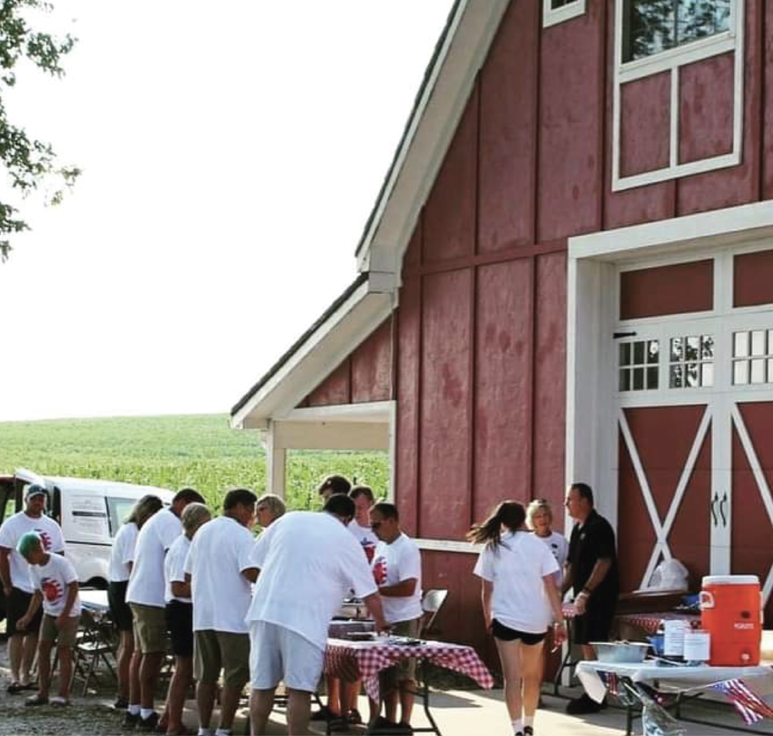 People gathered outside a red barn for a meal or event, with several tables covered with checkered tablecloths and food serving items, on a sunny day near green fields.