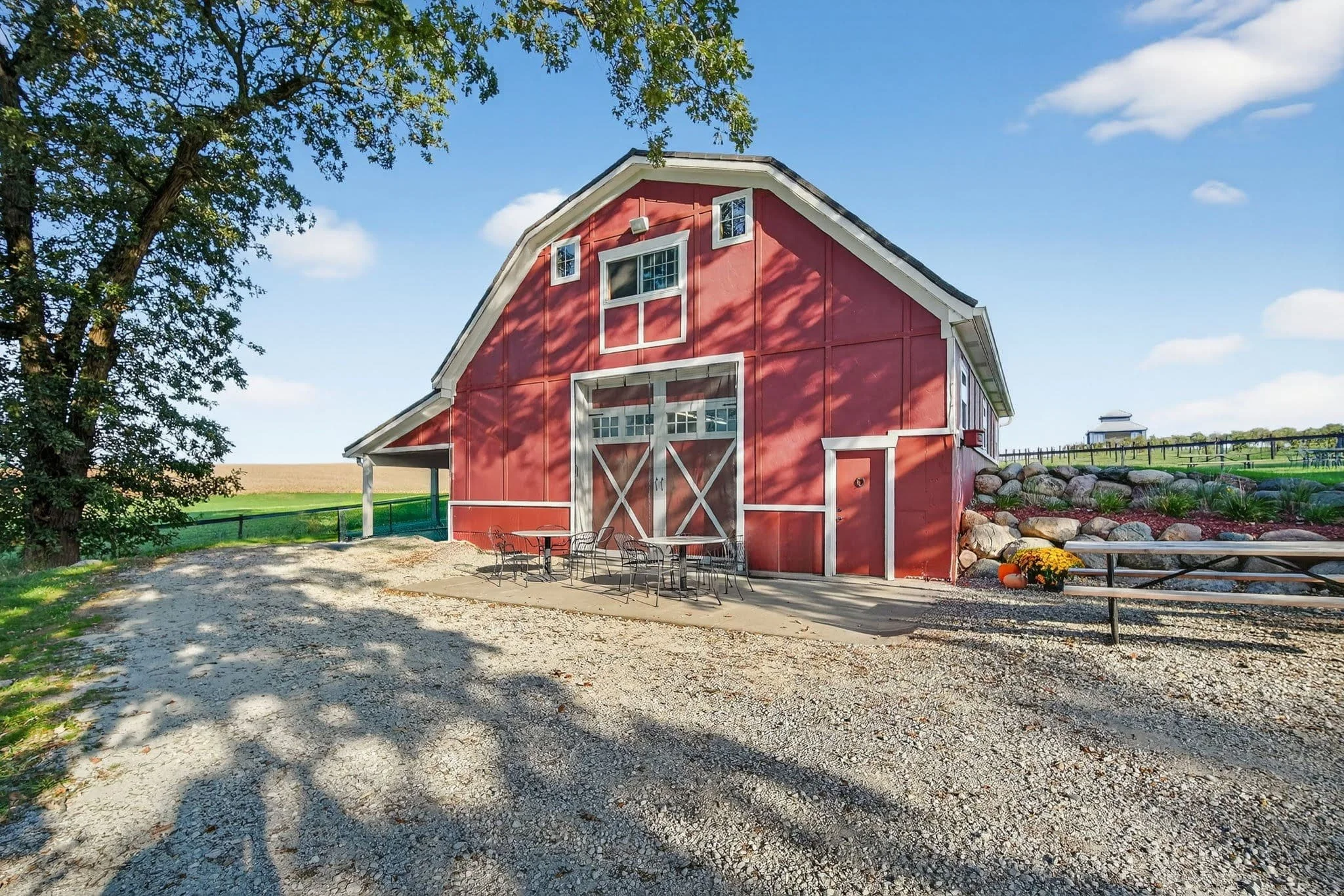 Red Barn event venue at Twin Vines on a sunny day, with patio tables and metal seating, and a gravel driveway. Perfect for event venue rentals in Panora, Iowa.