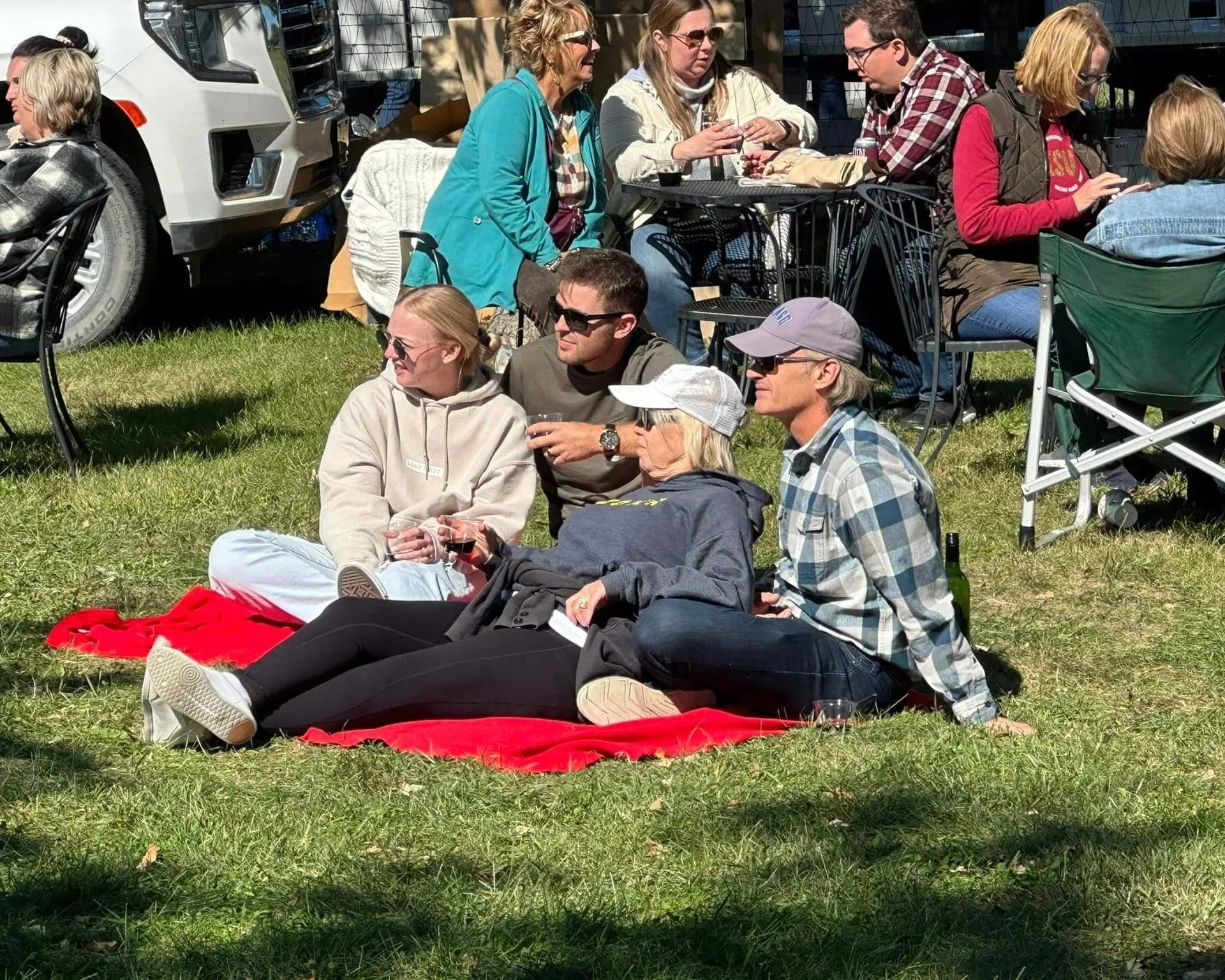 Family of four gathered on red blanket on lawn at Twin Vines with small plastic wine cups watching a live music performance at Twin Vines in Panora, Iowa.