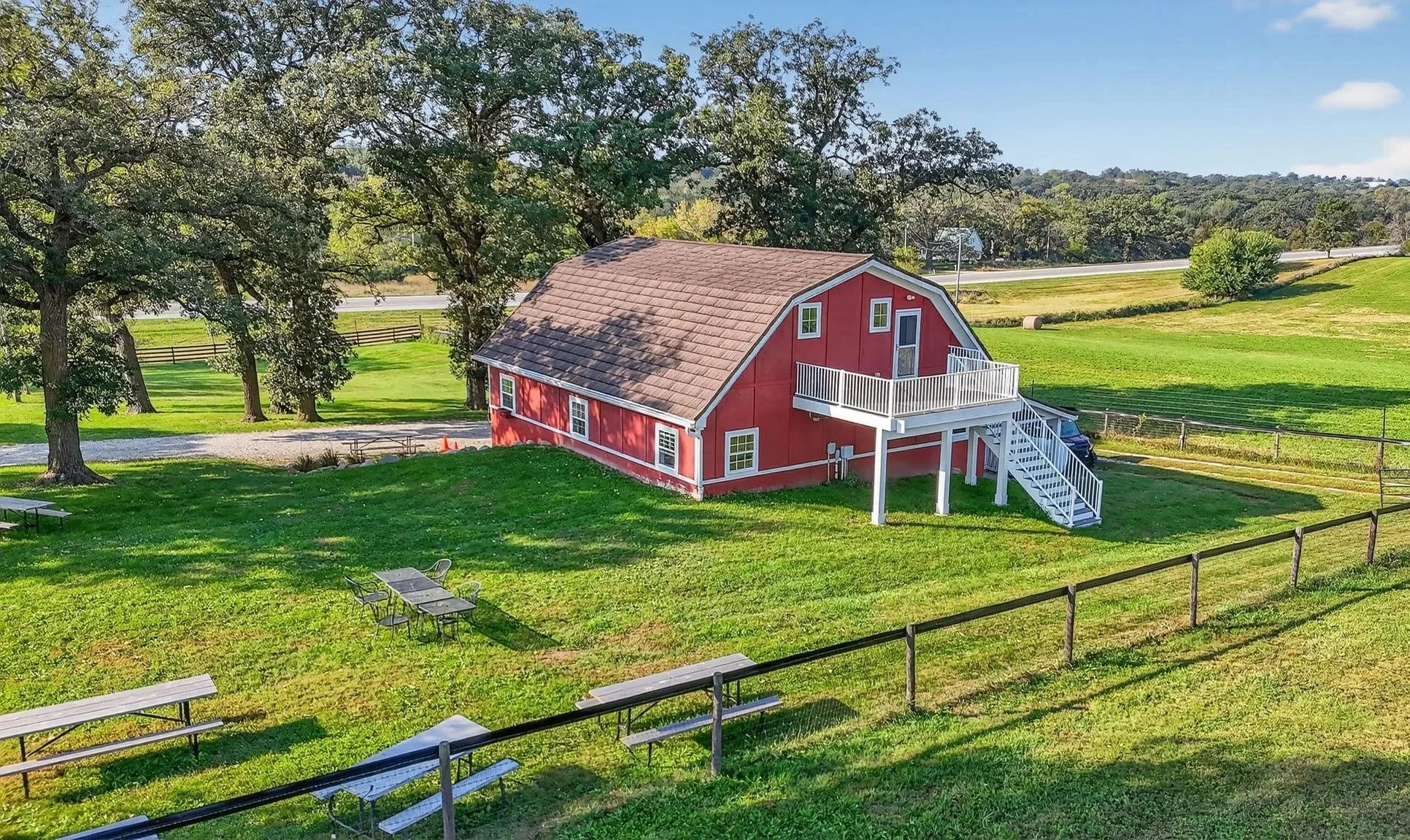 Red barn with white stairs leading to a second-floor balcony, surrounded by a green lawn and trees, with a scenic landscape of rolling hills and a blue sky in the background. Main event venue for private rentals at Twin Vines in Guthrie County.