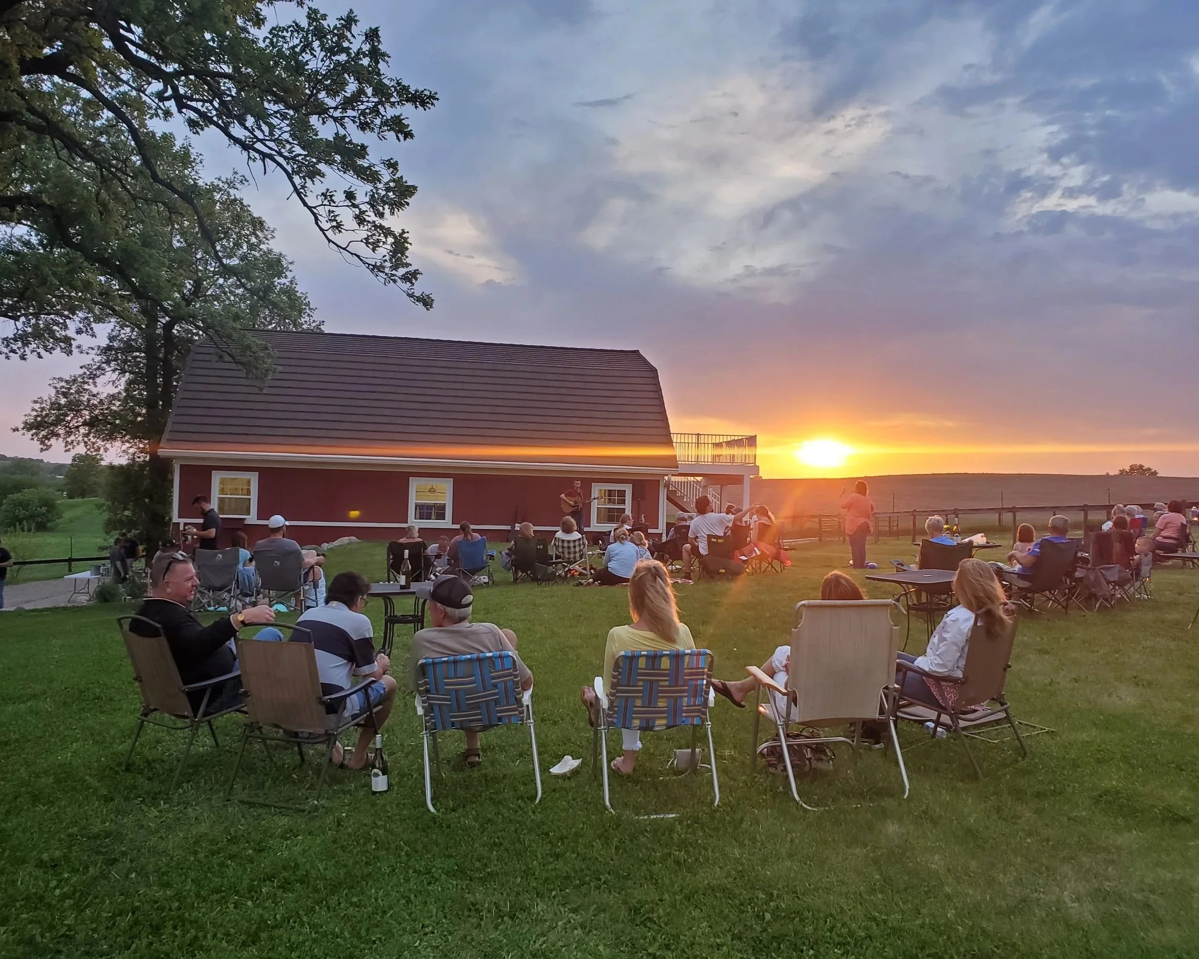 Community event/ live music performance at Twin Vines. 20+ people gathered on lawn behind red barn during sunset watching local musician sing and play guitar.