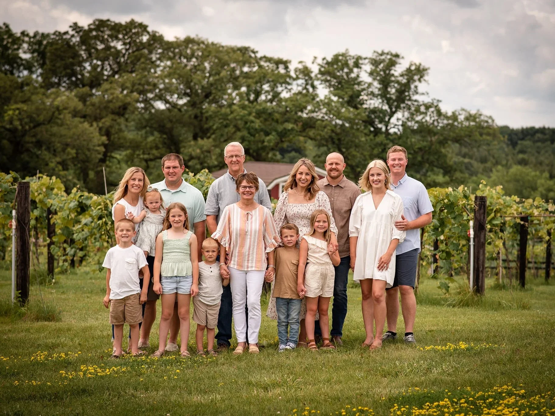 Family portrait taken during a private photography session at Twin Vines in Guthrie County, Iowa. Features 14 family members, both children and adults in front of vineyard ros standing atop dandelion-sprinkled grass.