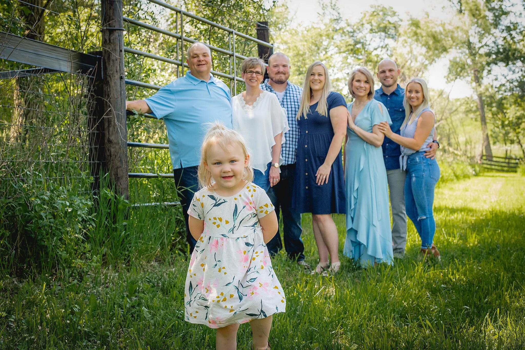 Family portrait of seven adults standing in grass near fence. Blonde little girl in forefront smiling at camera with hands behind her bacl. Taken during a private photography session at Twin Vines in Panora, IA.
