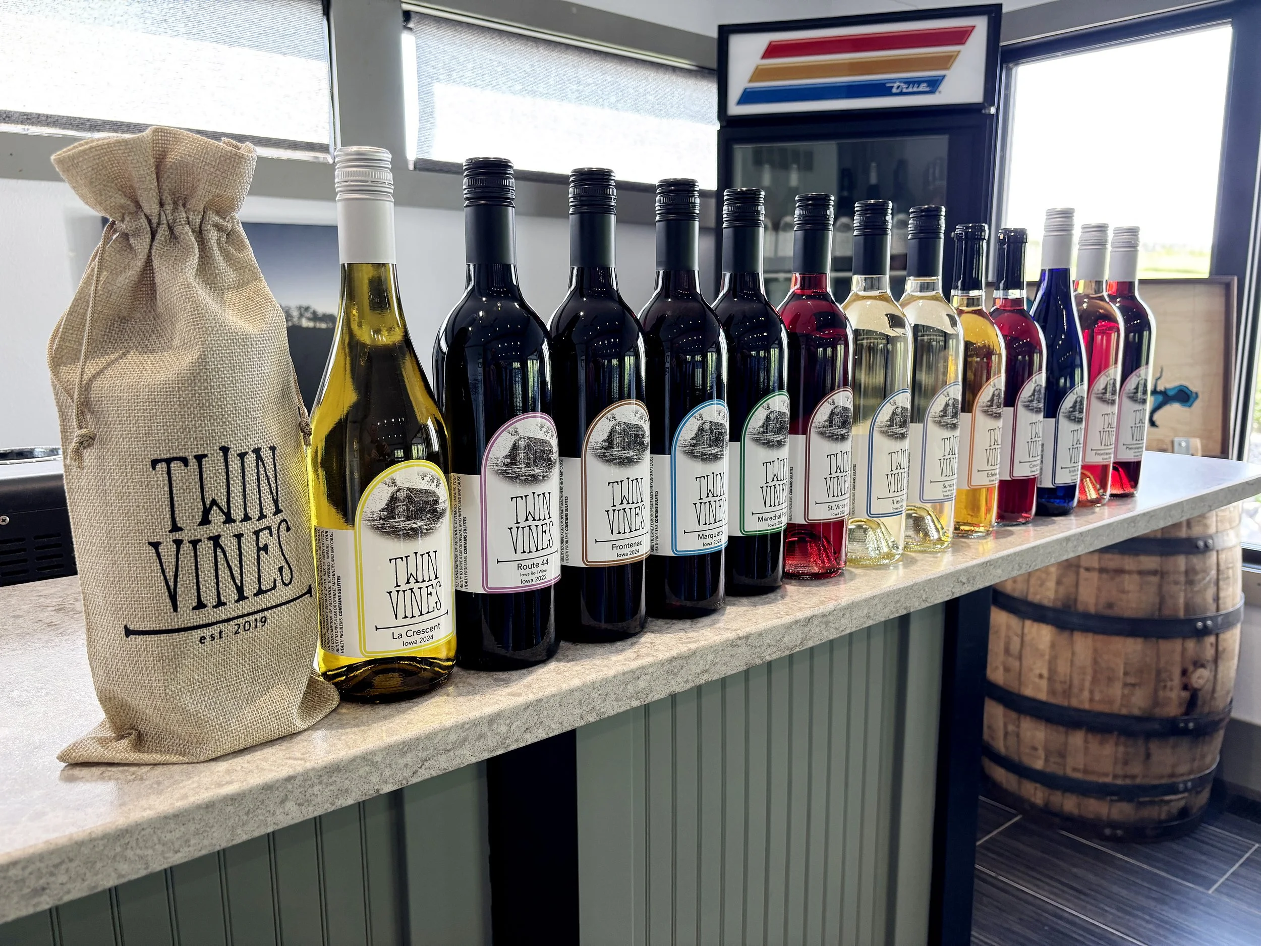 Twin Vines wines on a granite countertop, including dark and white bottles, next to a burlap sack with the Twin Vines logo, with a wine barrel and wine fridge in the background.