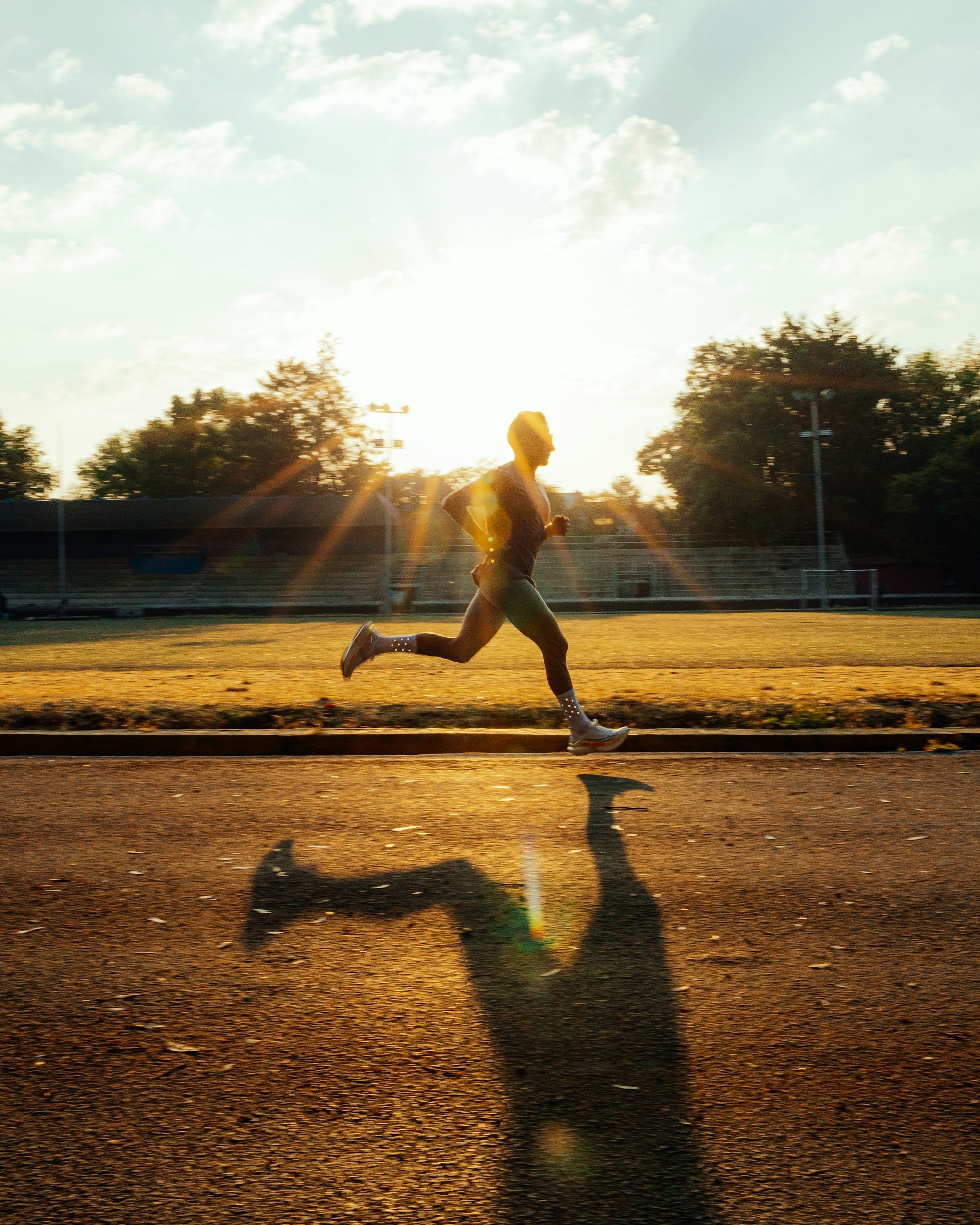 A person running outdoors on a track at sunrise or sunset, with the sun low on the horizon, trees in the background, and a clear sky with some clouds.