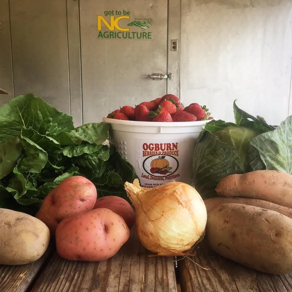 Basket of strawberries with lettuce, potatoes, an onion, and sweet potatoes on a wooden table, featuring "Ogburn Berries & Produce" label and "Got To Be NC Agriculture" sign in the background.