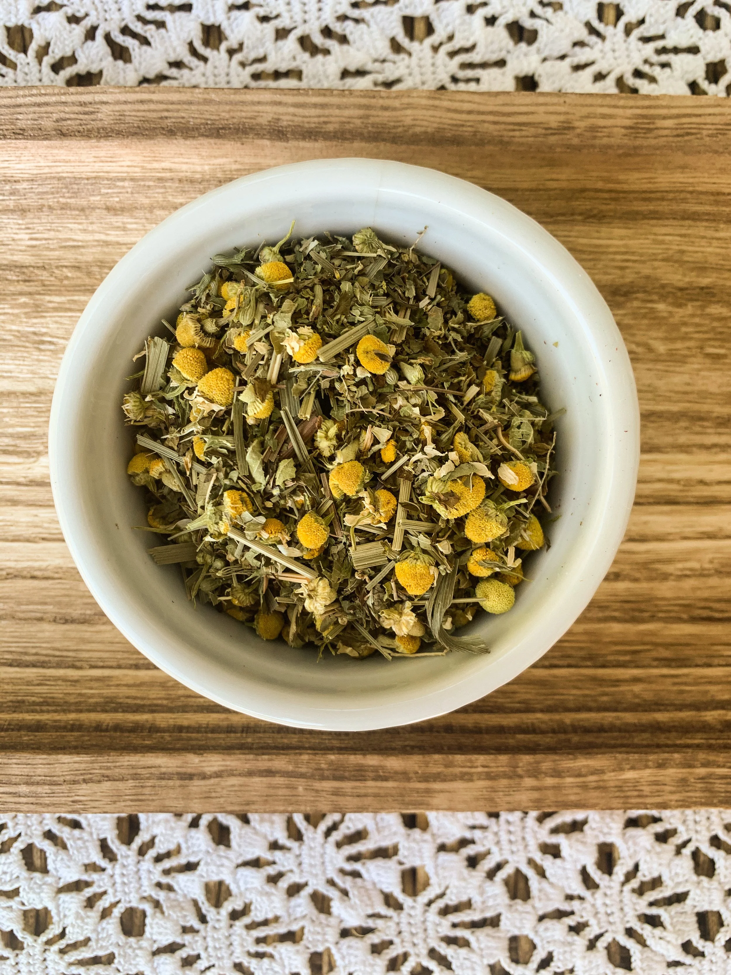 A white bowl containing dried herbal tea with chamomile flowers on a wooden surface.