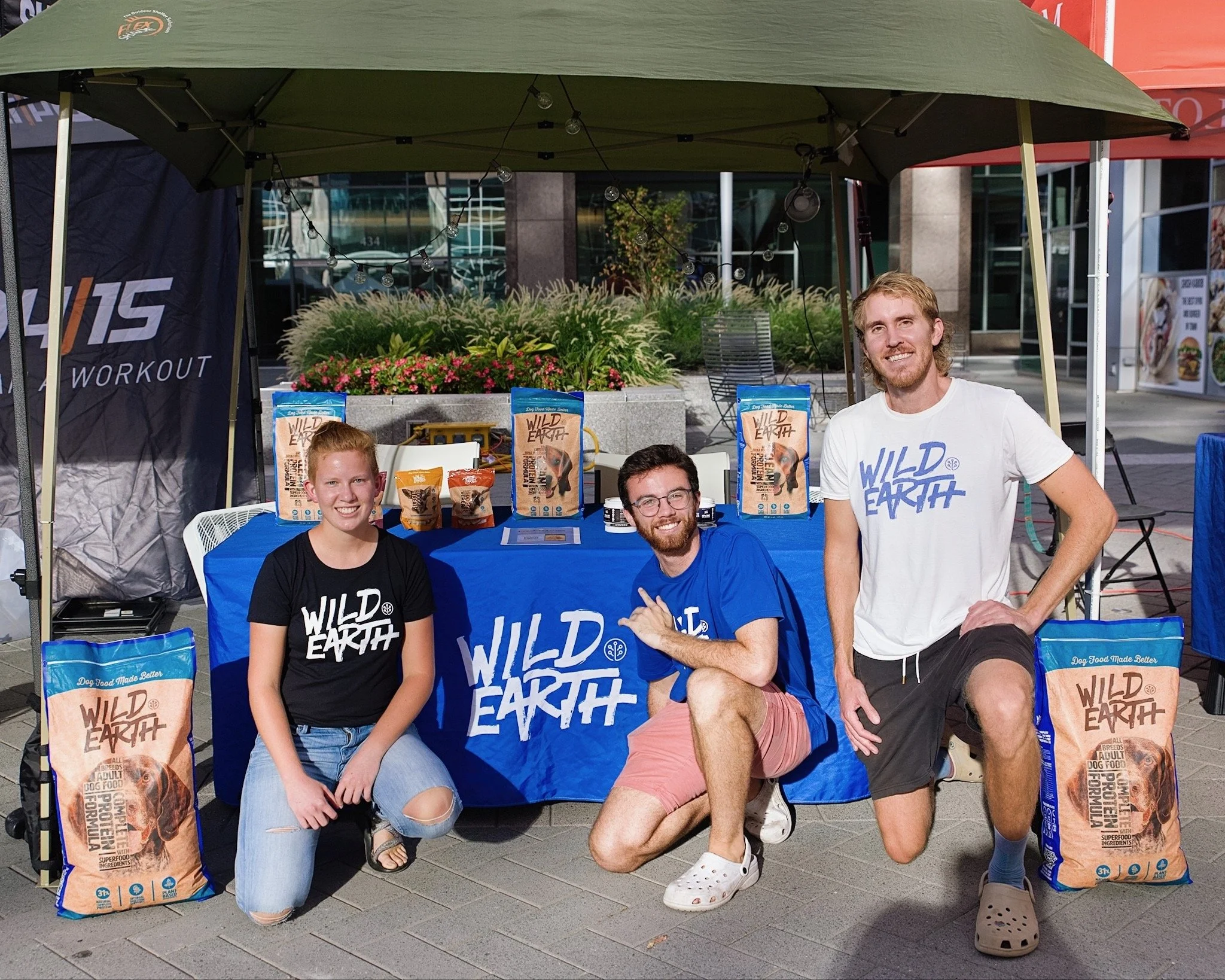 Three people wearing 'Wild Earth' shirts posing at a booth with pet food packages on display under a canopy.