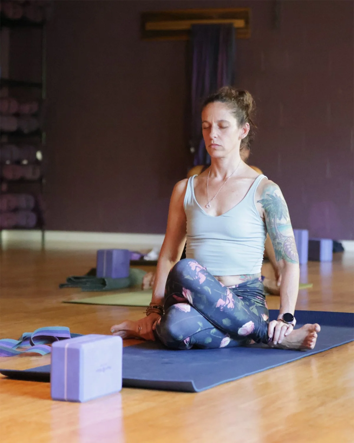 xWoman seated on a yoga mat in a gentle twist with eyes closed in a calm yoga studio.