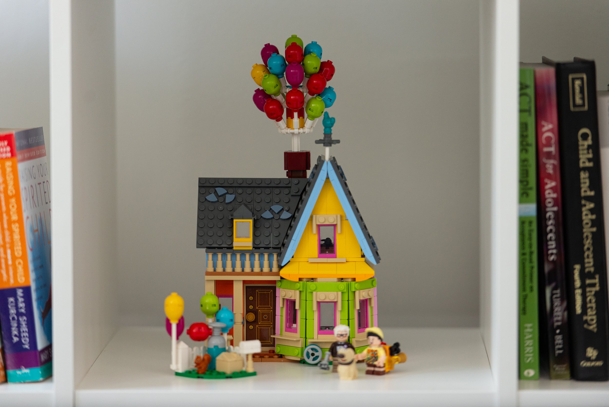 A LEGO house with colorful balloons on the roof and a small LEGO garden and characters in front, placed on a white shelf between books.