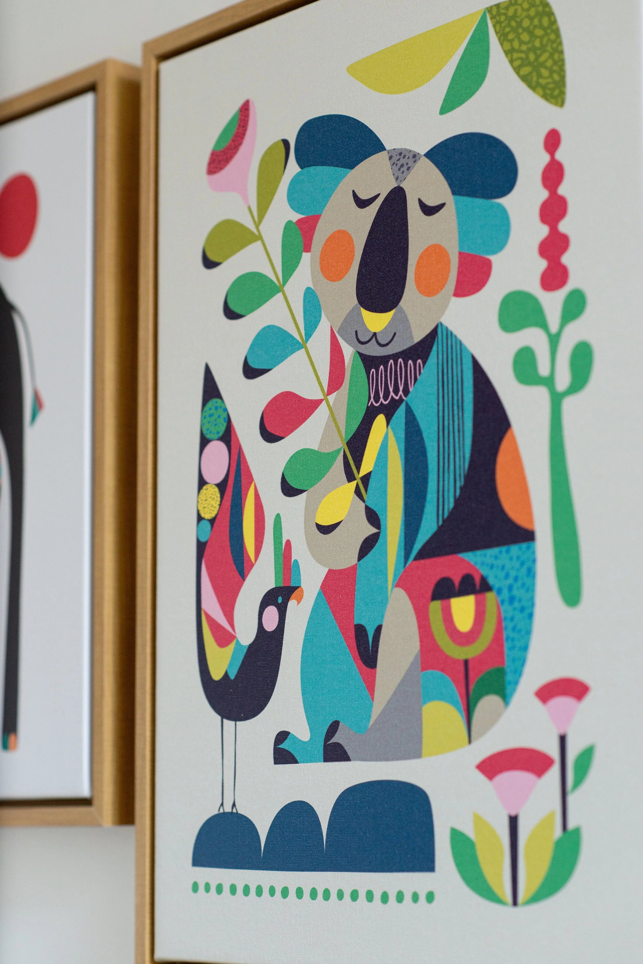 Colorful abstract illustration of a seated koala holding a branch with leaves, accompanied by a black bird, set within a wooden frame.