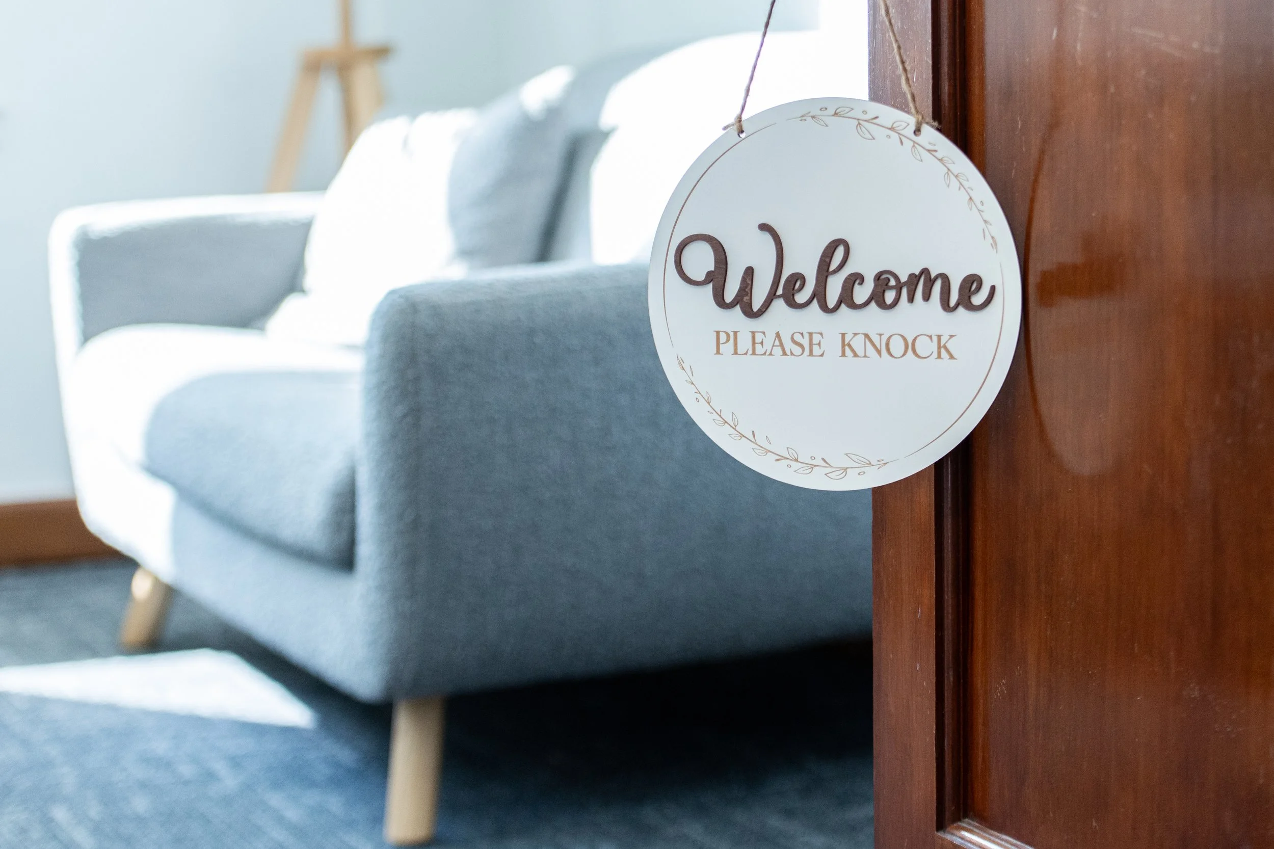 A round welcome sign hanging on a wooden door with the words "Welcome, Please Knock" written on it, near a light gray sofa in the entryway to Dr. Wade's Raleigh office.