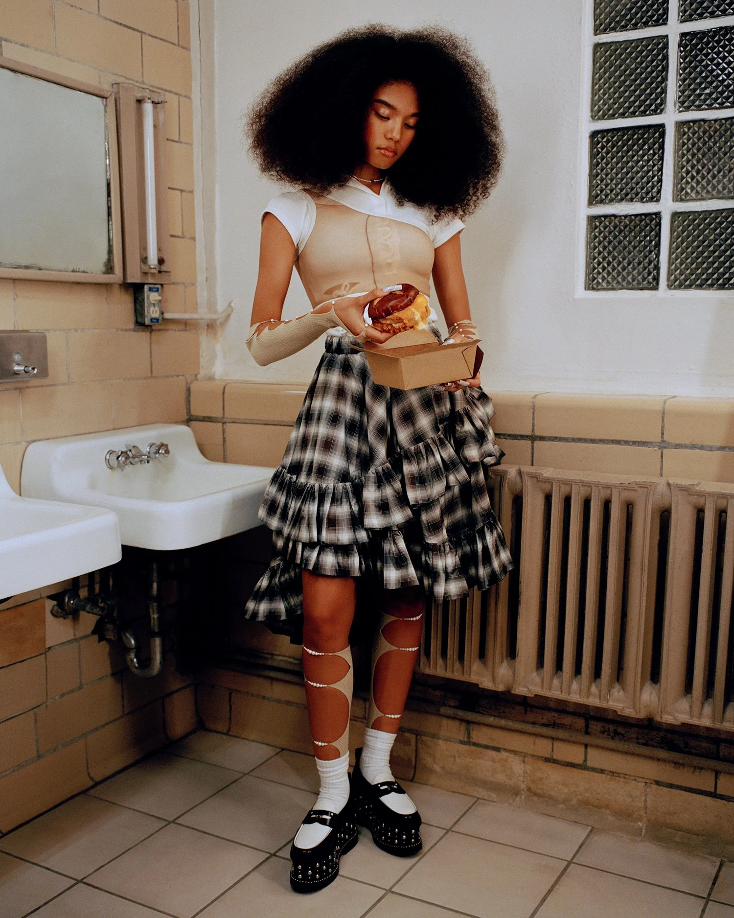 A young woman with large curly hair stands in a institutional-looking room holding food in a takeout box. She wears a layered outfit including a white top, beige and see-through arm sleeves, a ruffled plaid skirt, eccentric patterned tights, and chunky black and white shoes.