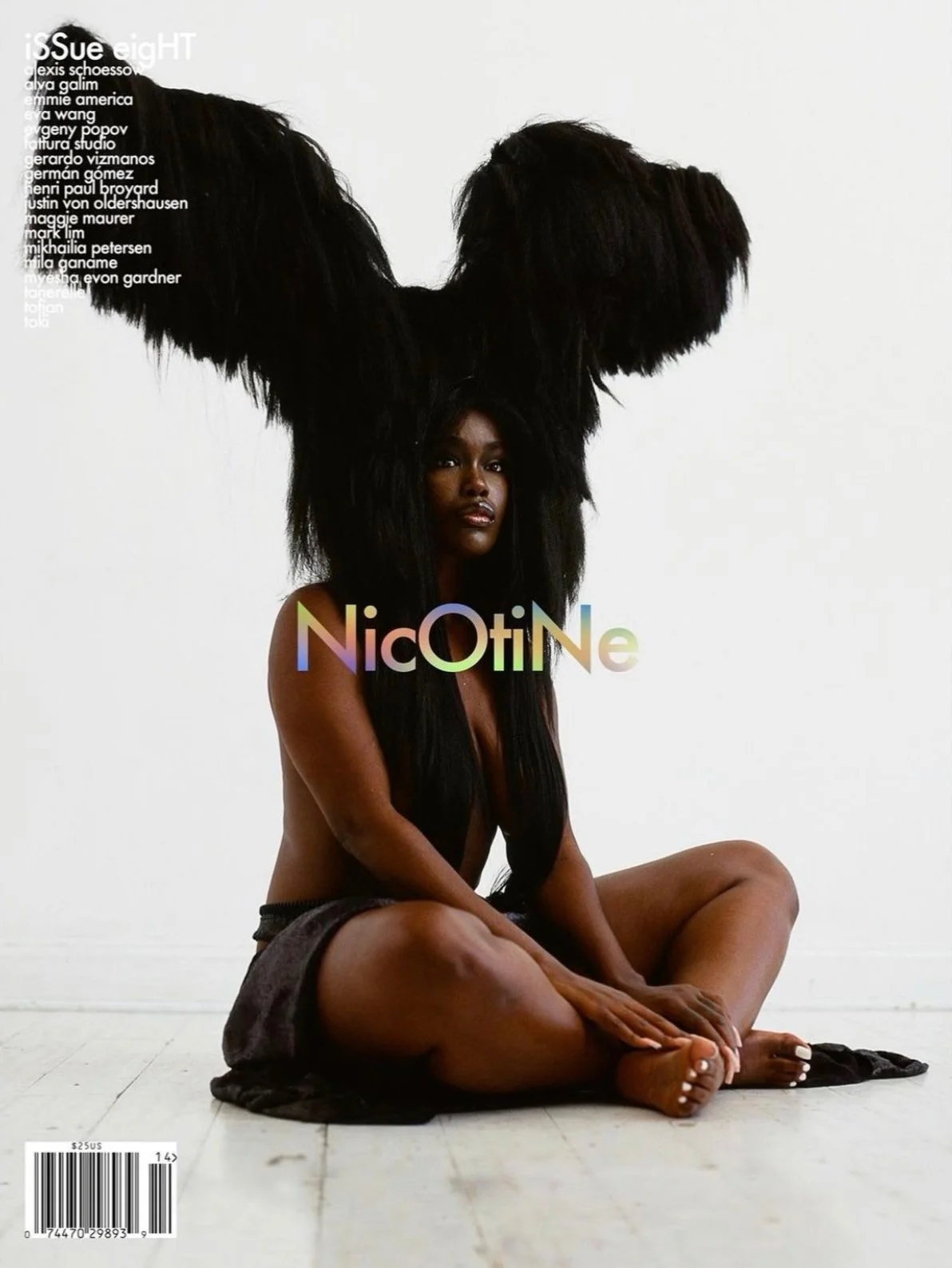 A woman with long dark hair sitting cross-legged on the floor against a plain white background, with a large black feathered headpiece behind her. The magazine cover displays the title 'NicOTiNe' in colorful text at the center.